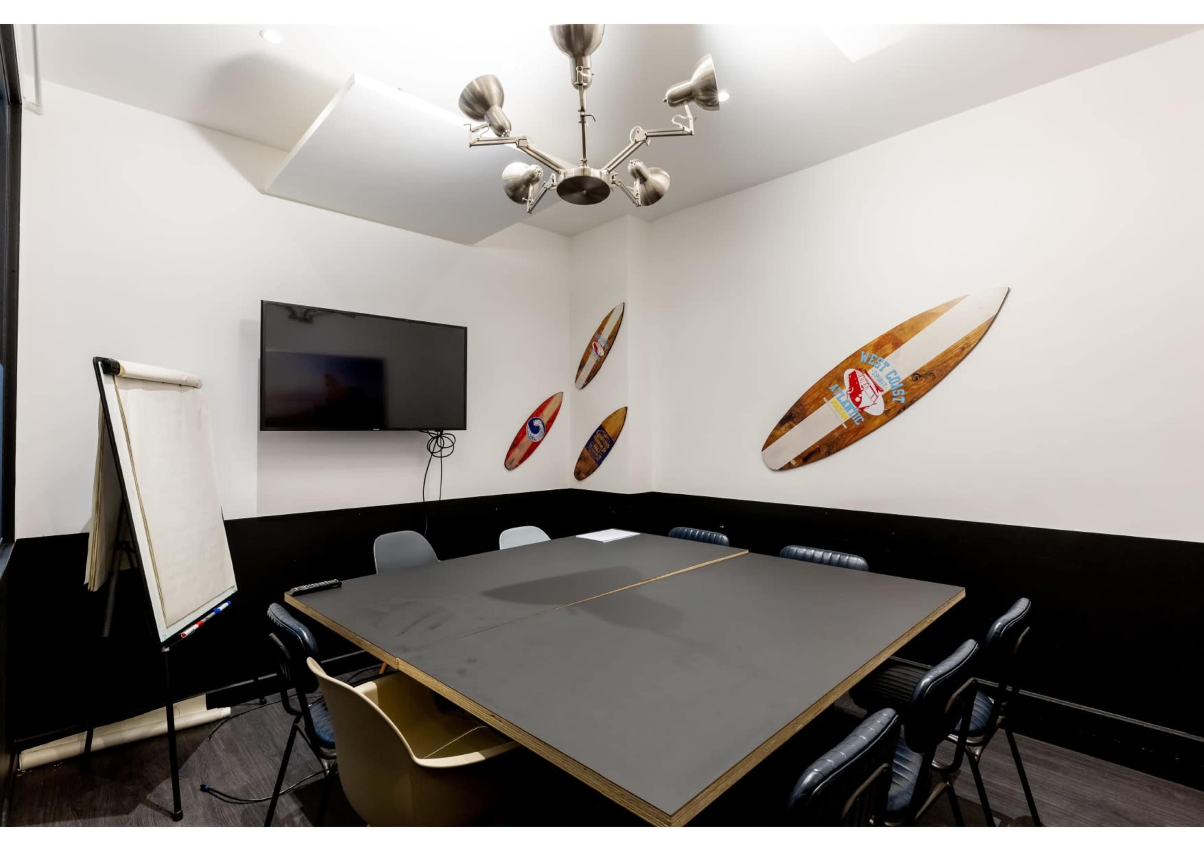 A modern conference room with a large black table, seating around it, a wall-mounted television, and surfboards displayed on the walls.