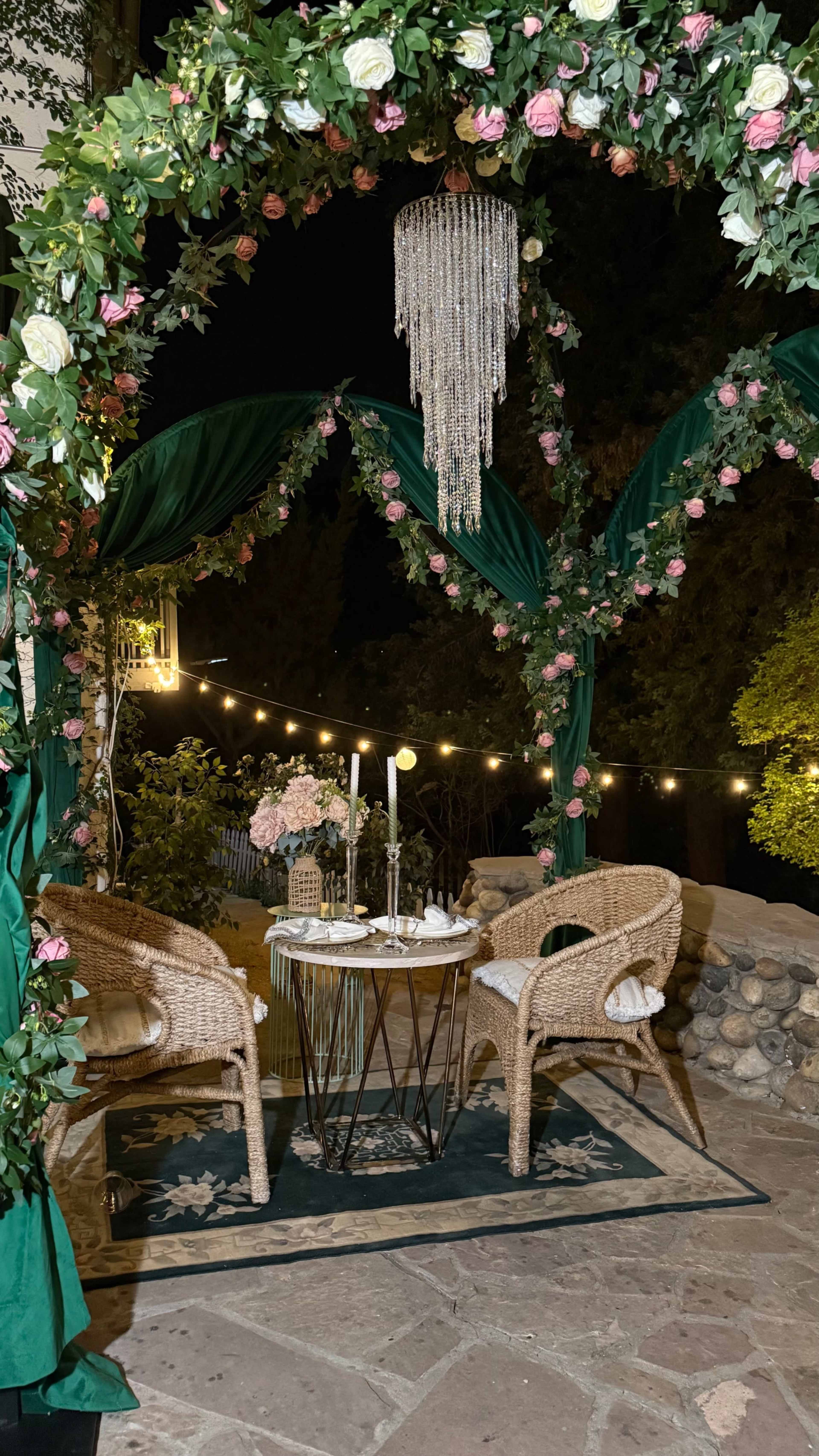 A cozy outdoor dining setup features a round table with candles, two wicker chairs, and an arch adorned with greenery and flowers beneath a sparkling chandelier.
