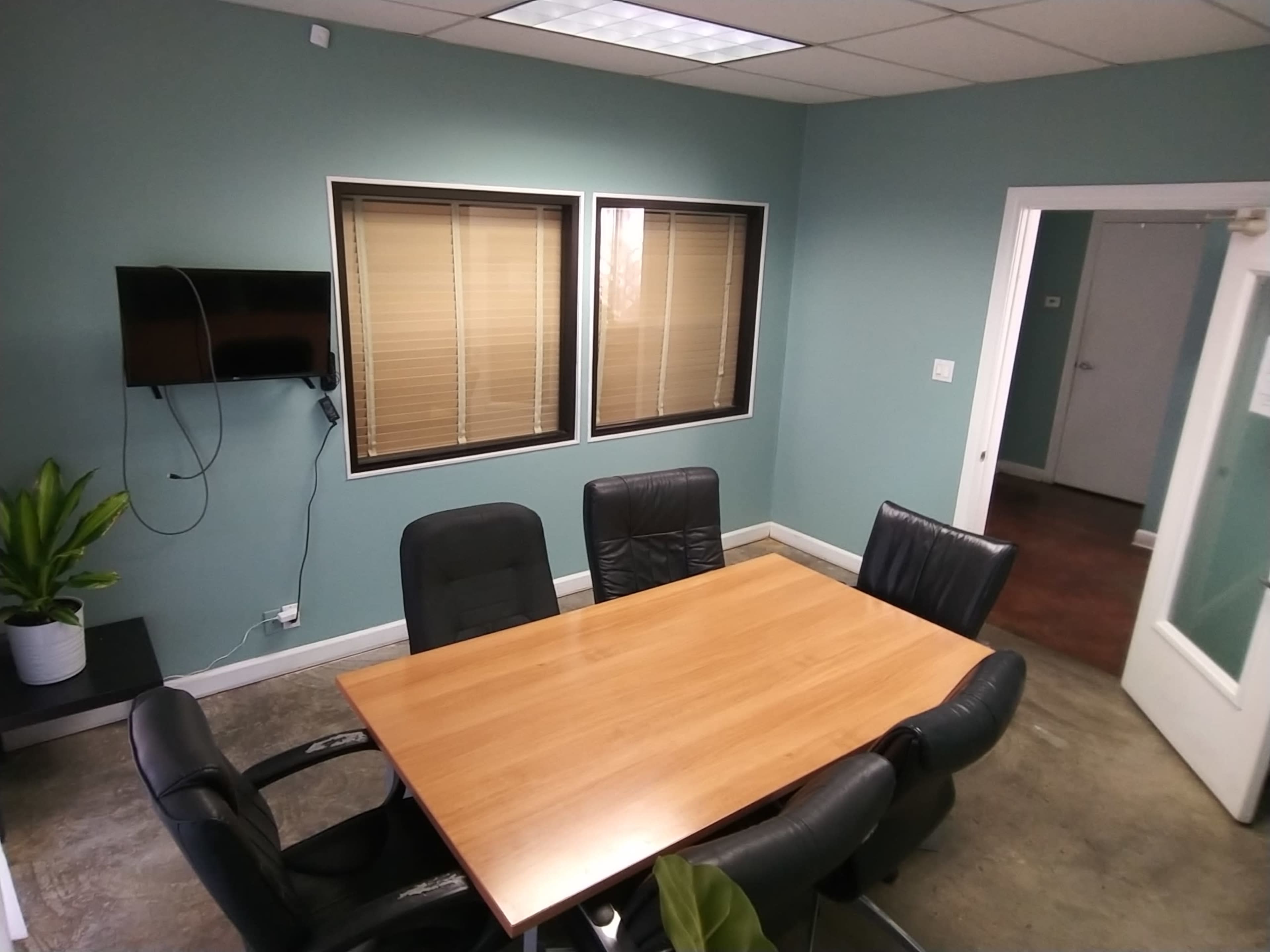 A meeting room with a rectangular wooden table surrounded by six black chairs, two windows with blinds, a wall-mounted TV, and a potted plant in the corner.