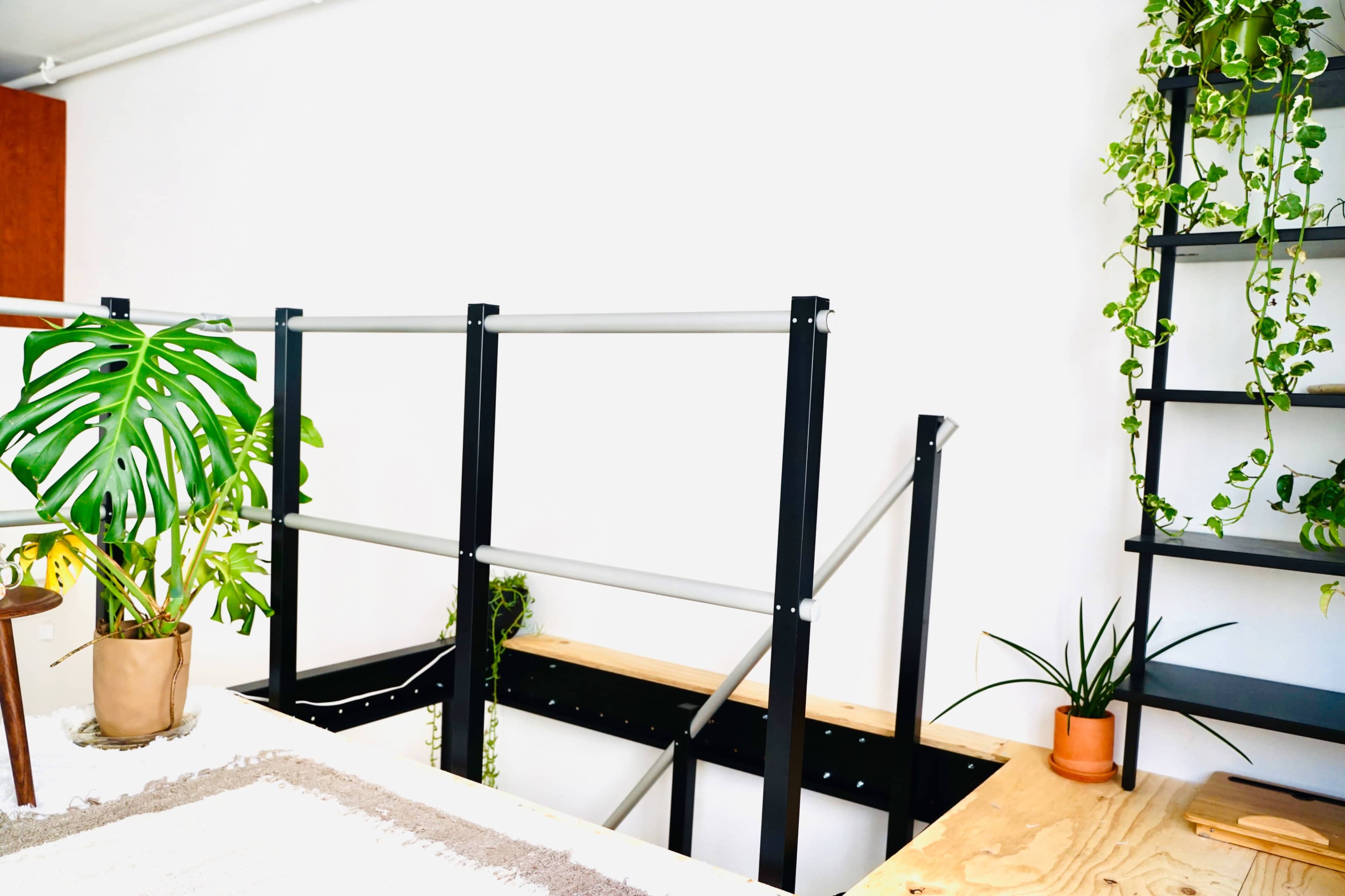 The image shows a minimalistic indoor space featuring a staircase with metal railings, wooden platforms, and various potted plants along the walls.