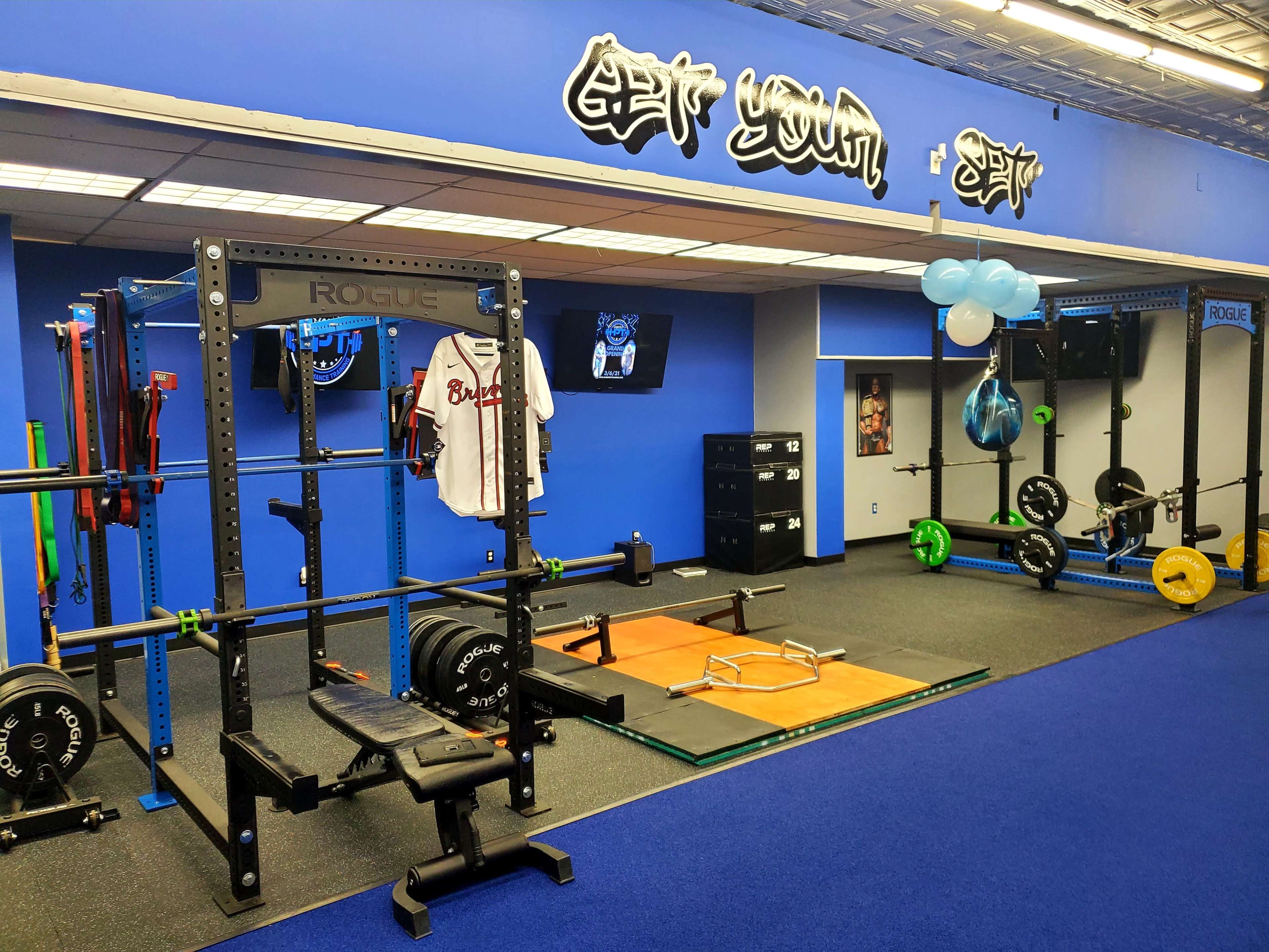 A gym interior with weightlifting equipment, a barbell on a weightlifting platform, and decorations including balloons and a sports jersey on display.