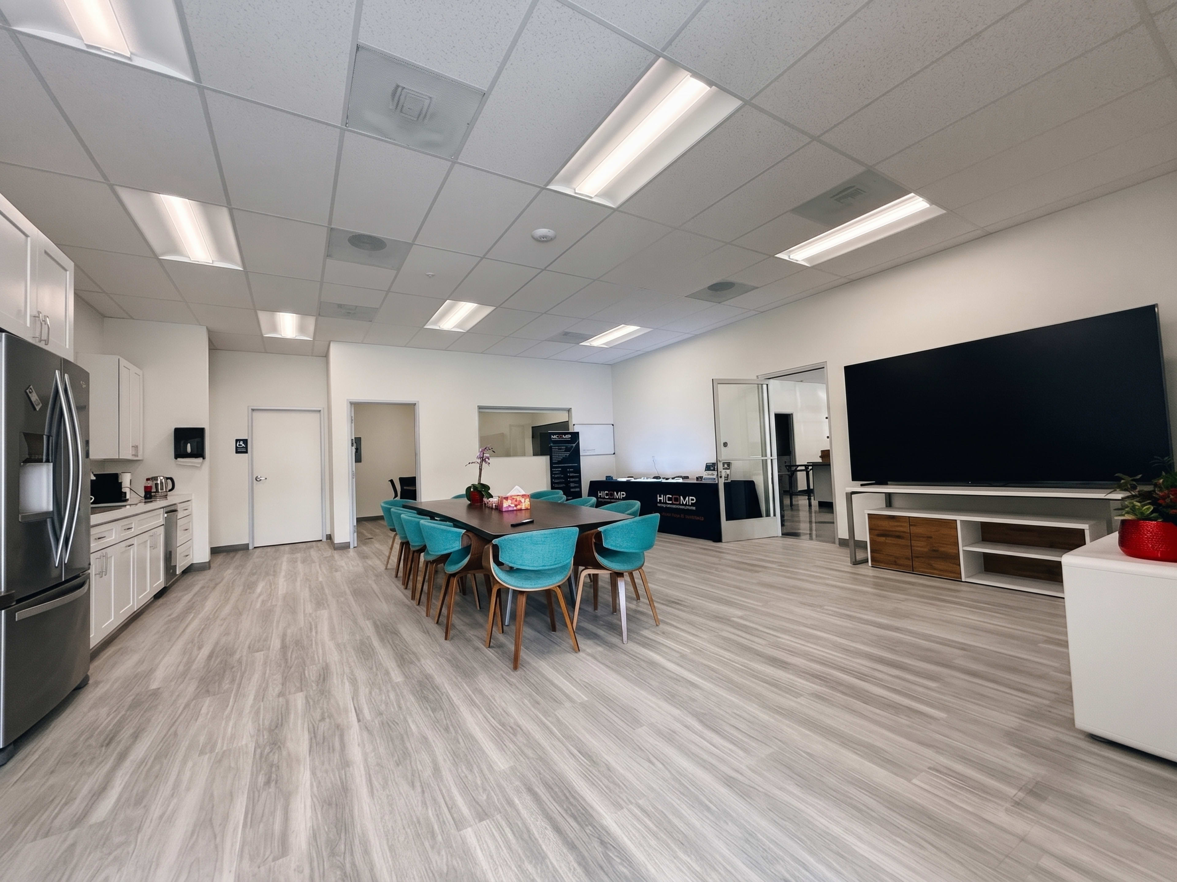 The image shows a modern office kitchen area with a dining table, chairs, a refrigerator, and a large television.