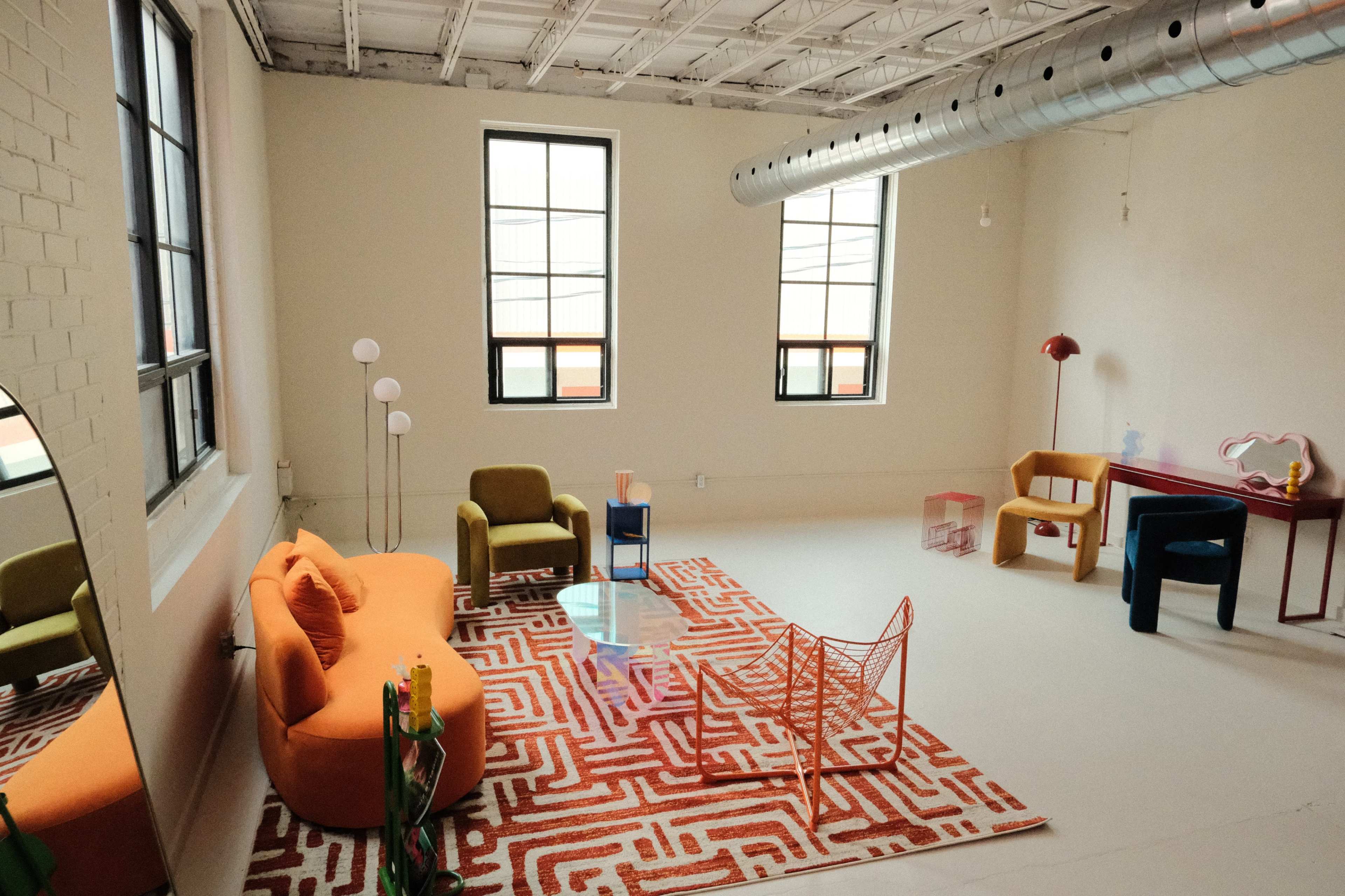 The image shows a modern, open living space with colorful furniture, a patterned rug, and large windows.