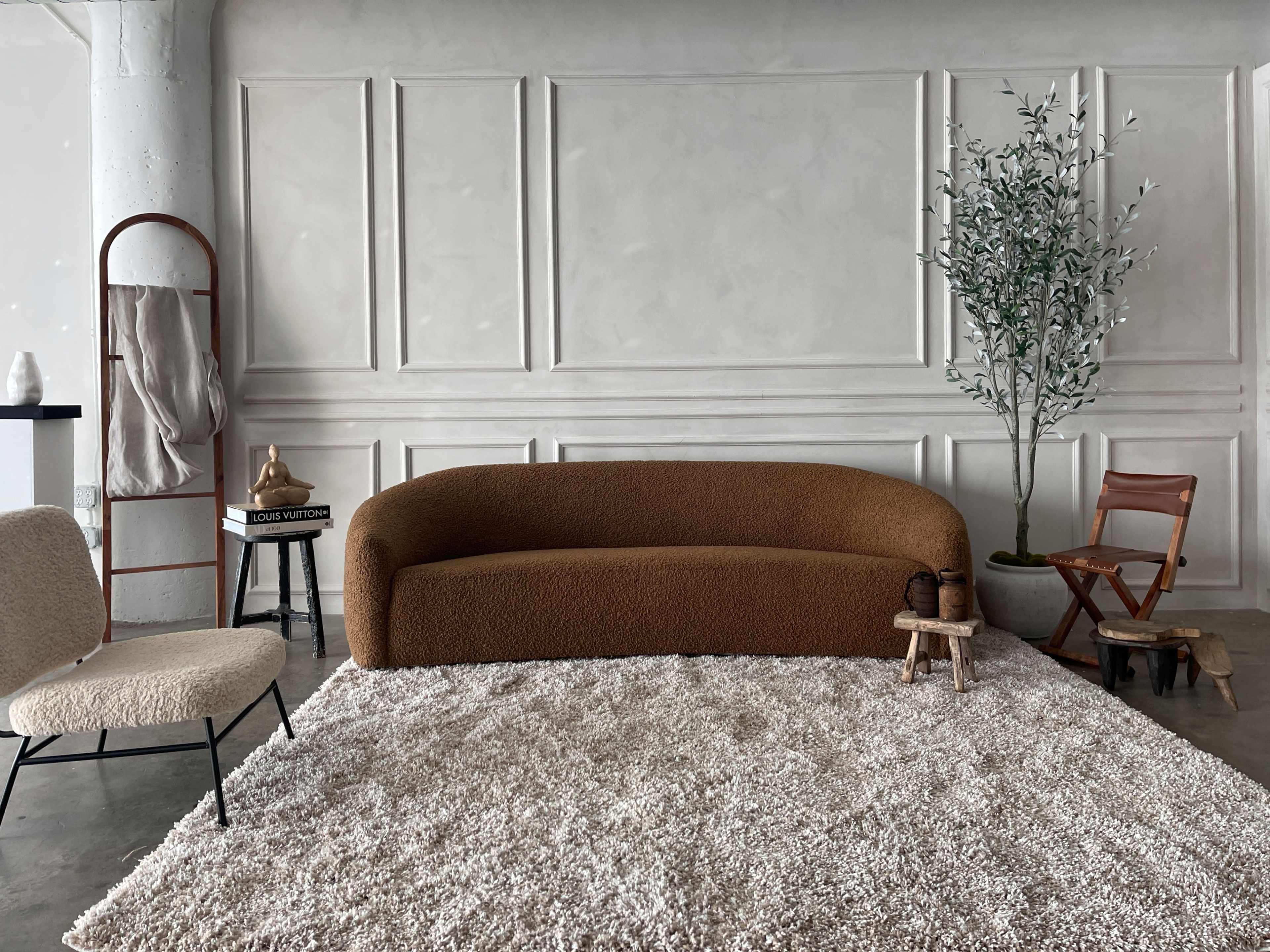 The scene features a brown, rounded sofa placed on a plush, light-colored rug, with a small wooden table, a chair, and a potted plant nearby, set against a textured, off-white wall.