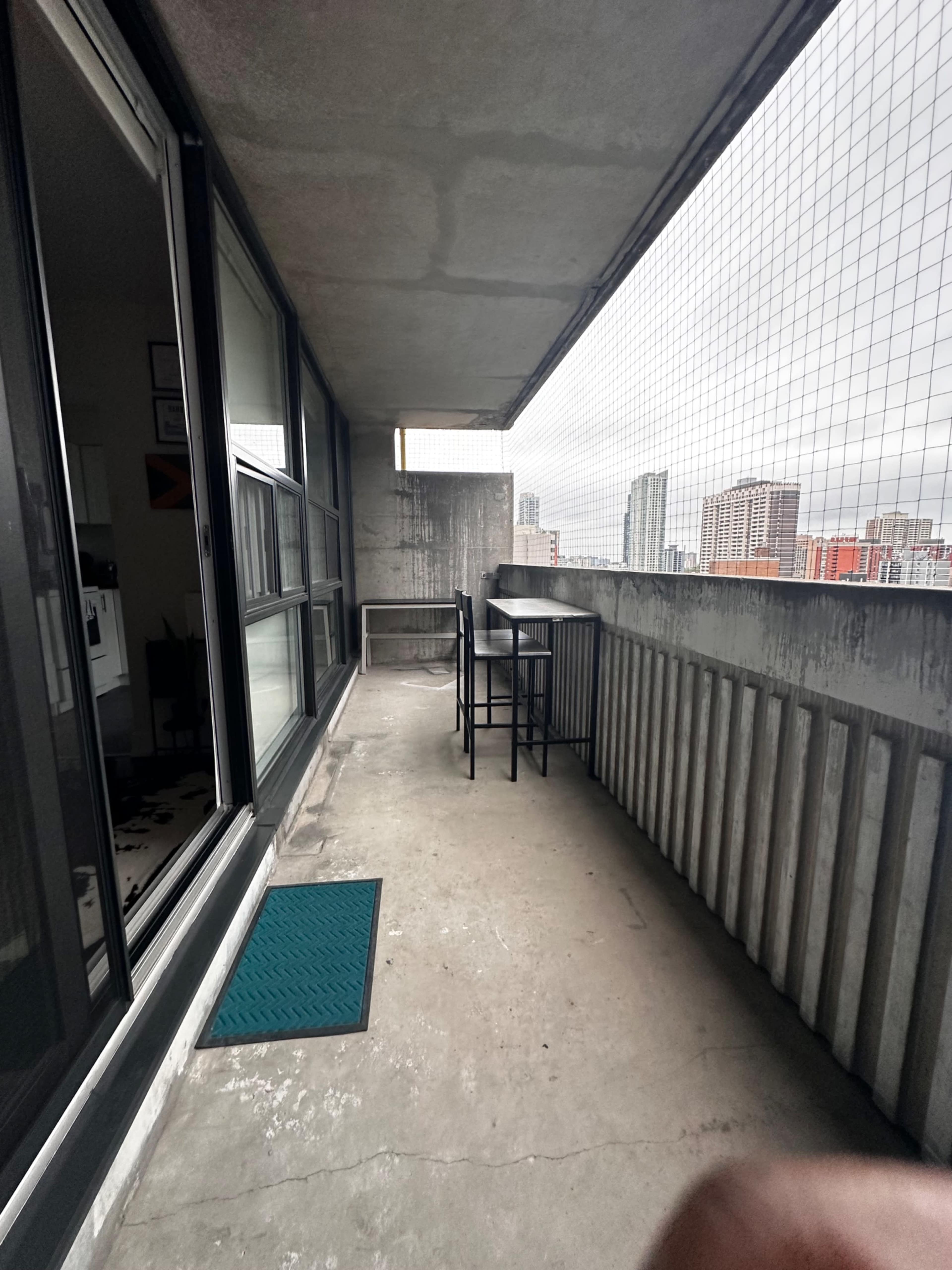 A narrow balcony features a small table and high chairs, overlooking a cityscape with tall buildings.