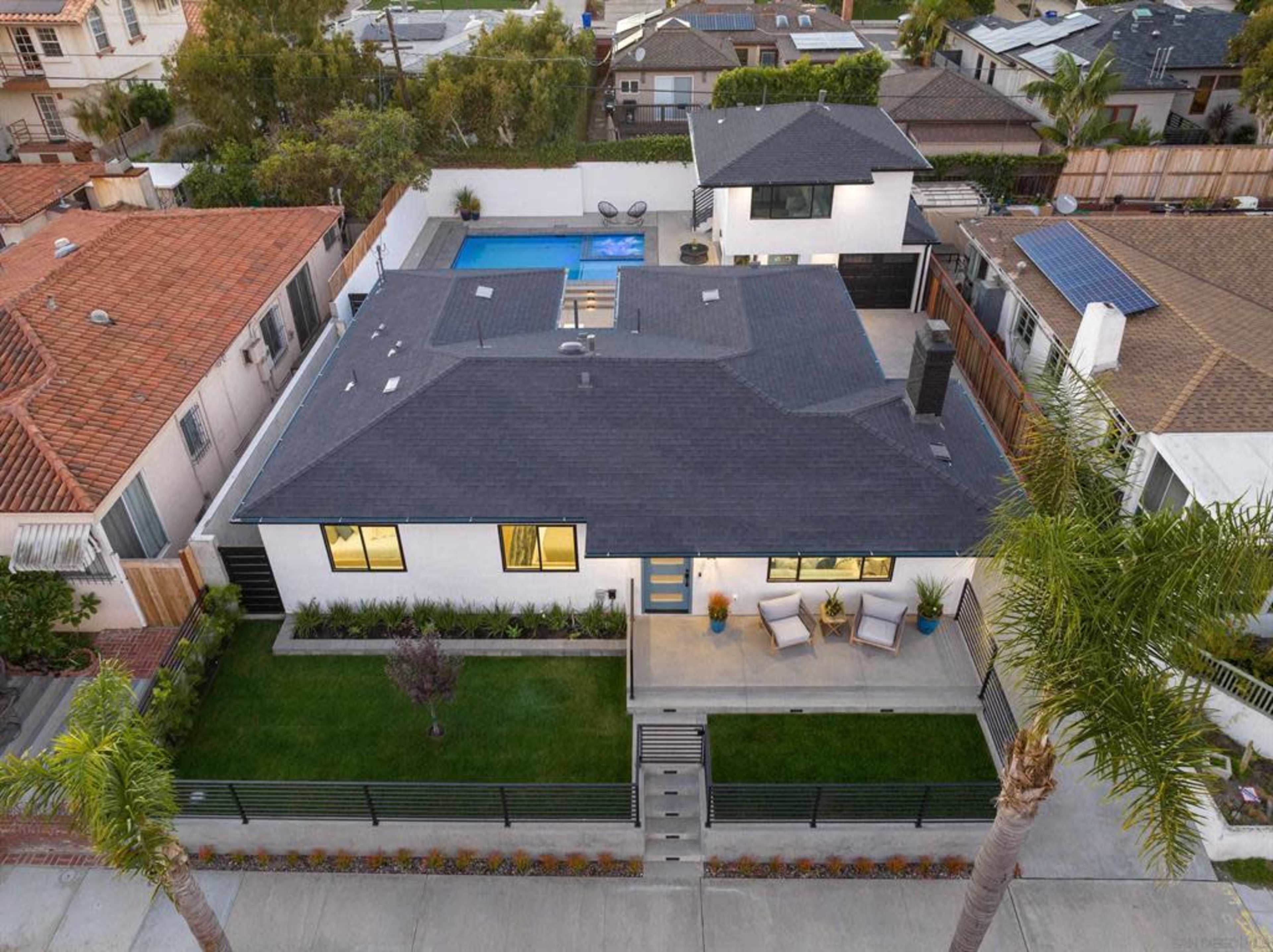 A modern house with a swimming pool and outdoor seating area, surrounded by neatly landscaped green space and a black fence.
