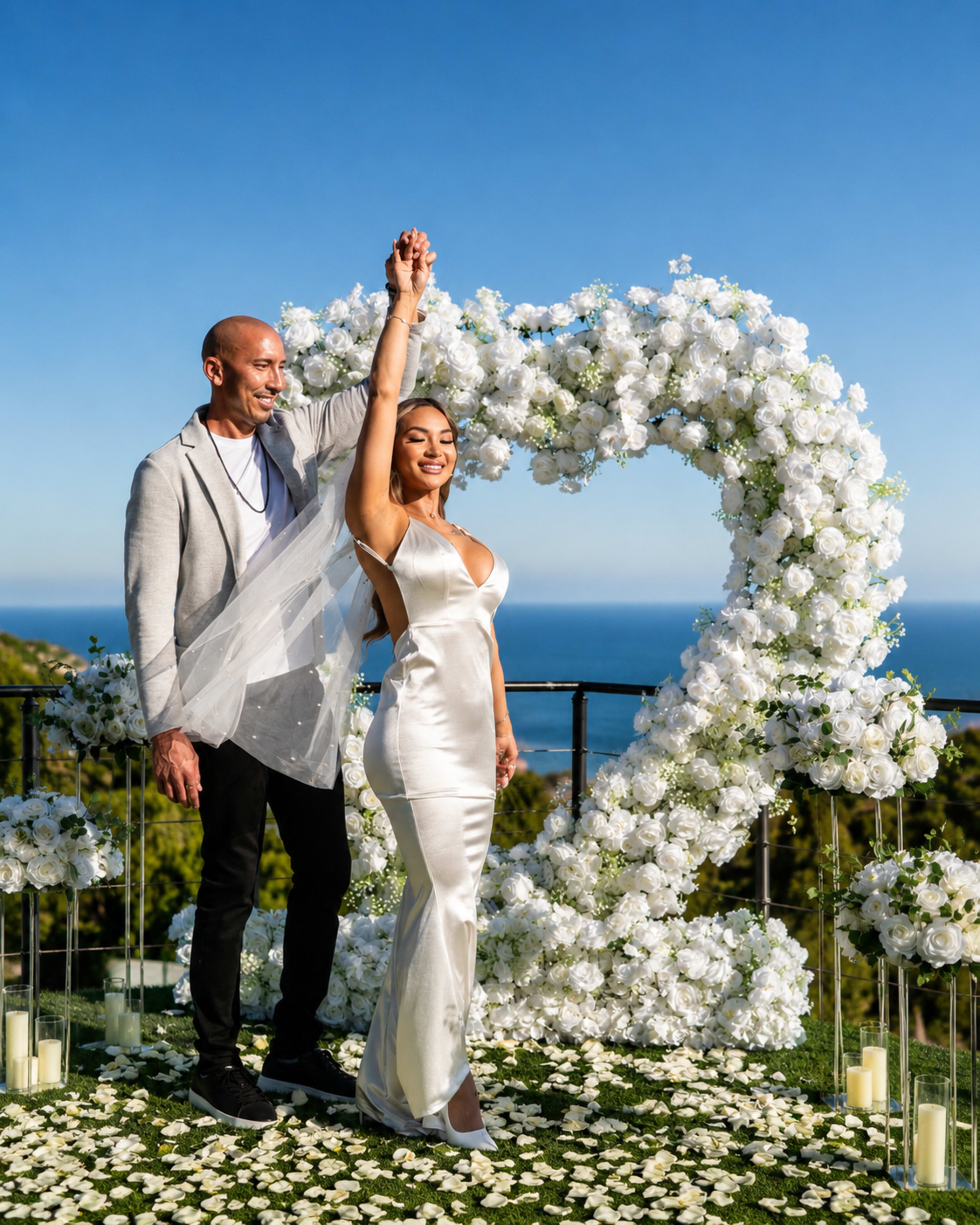 A couple poses joyfully in front of a heart-shaped floral arrangement on a balcony overlooking the ocean.