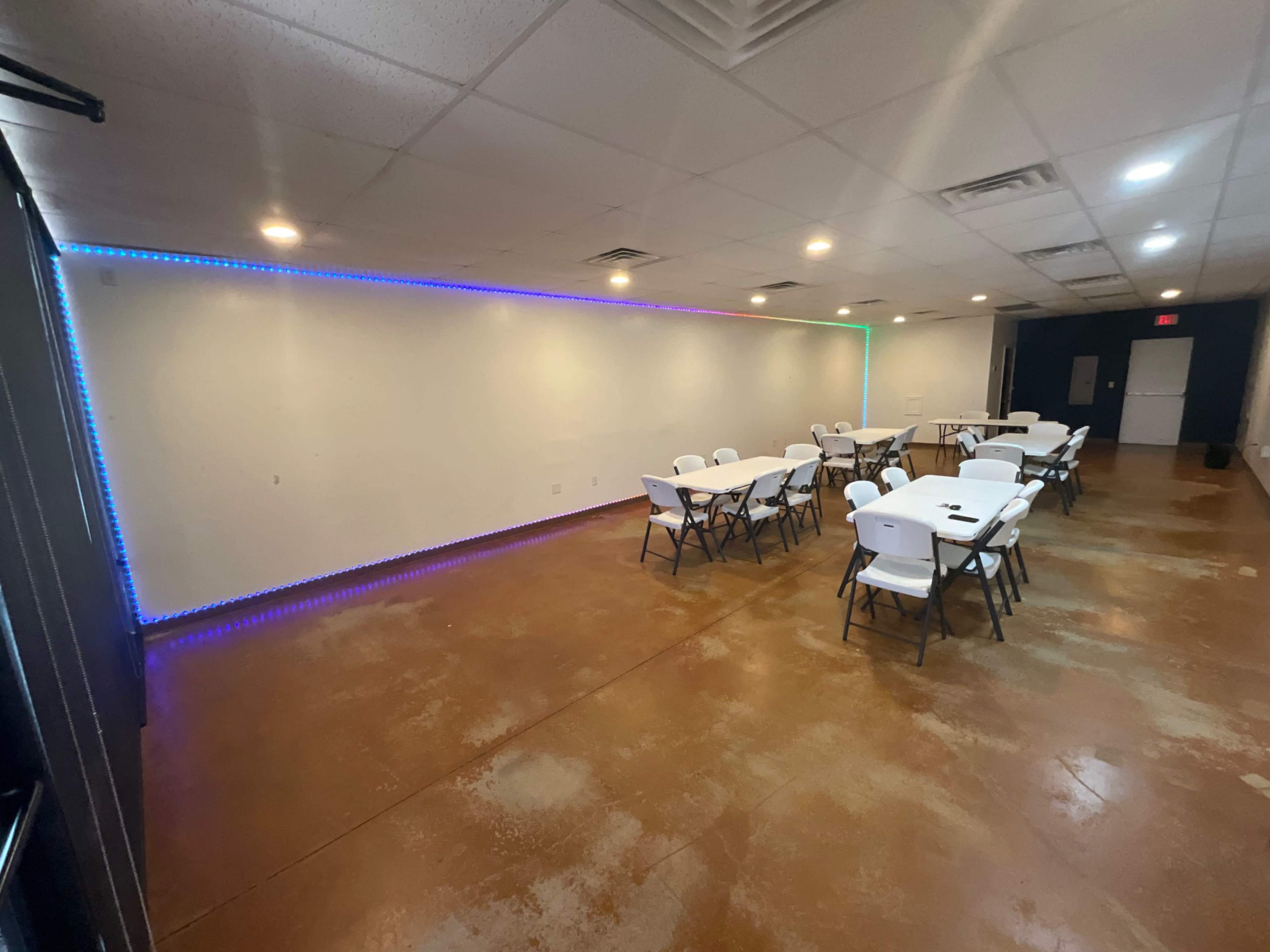 The image shows a spacious room with several white tables and chairs arranged, and colorful LED lights running along one wall.
