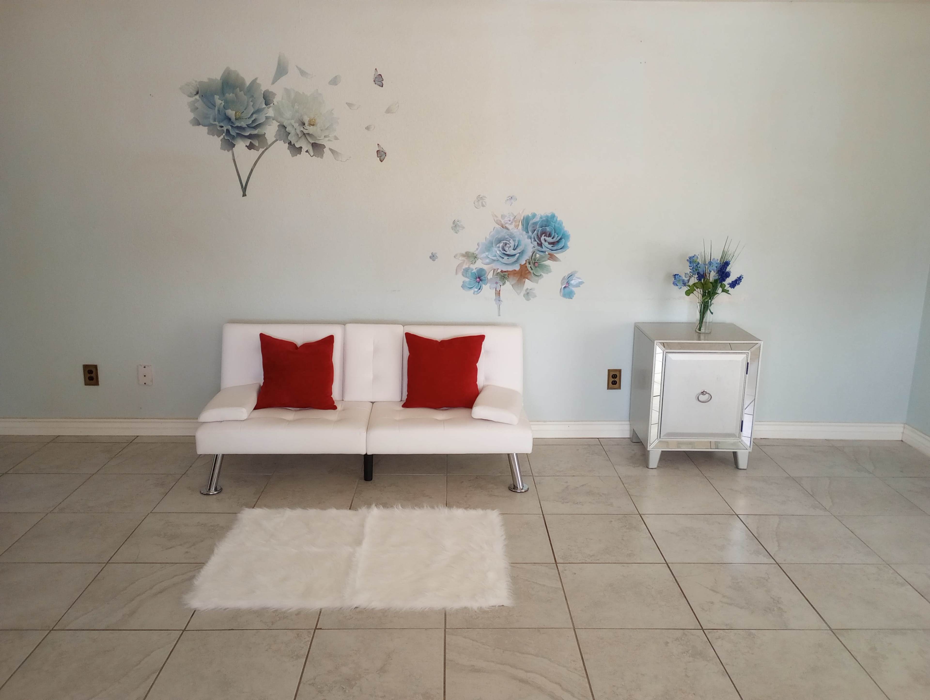 A minimalist living area featuring a white couch with red cushions, a small mirrored side table, and floral wall decor on a light blue wall.