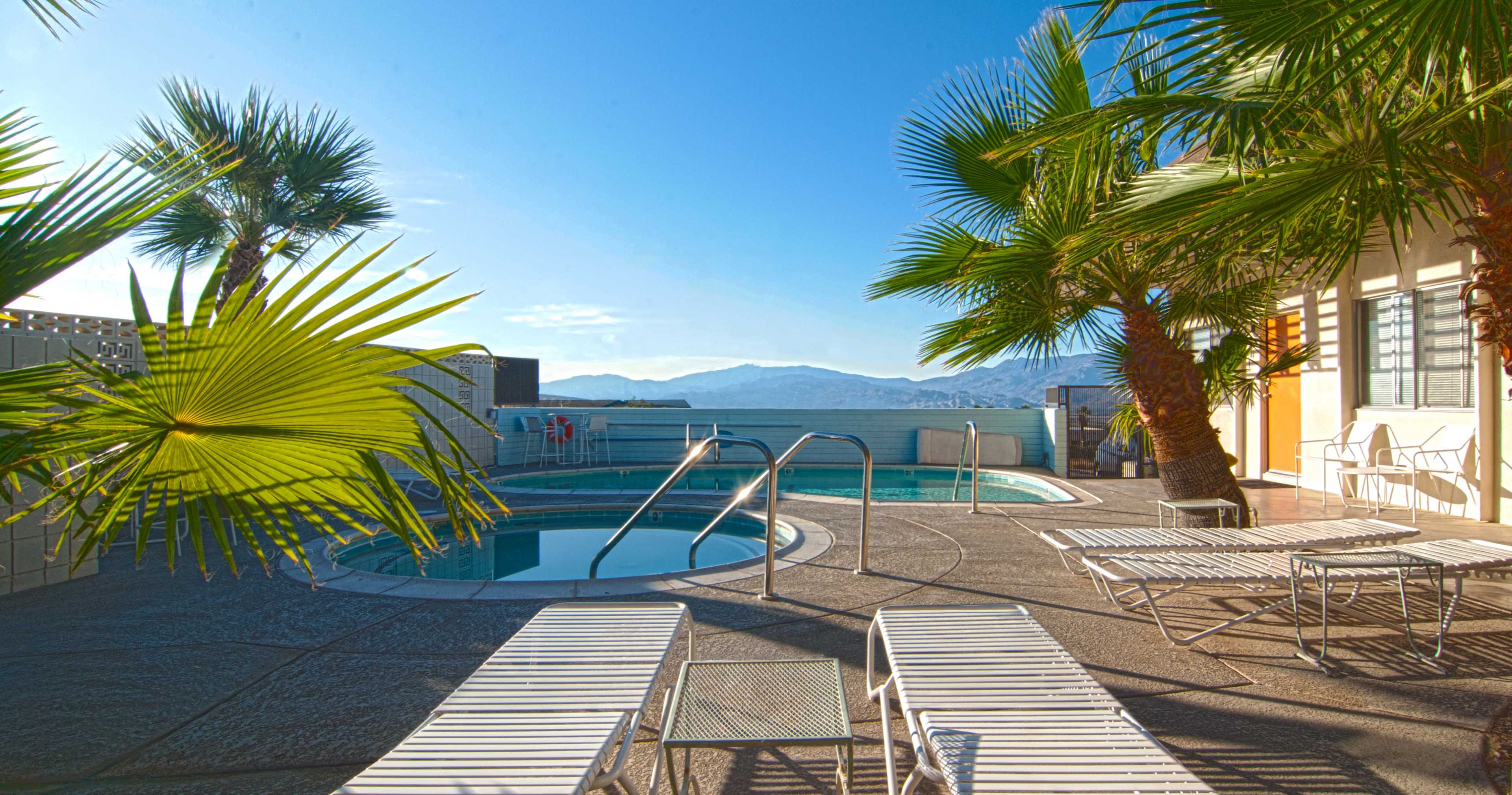 The scene features a swimming pool area with lounge chairs and palm trees, set against a backdrop of mountains and a clear blue sky.