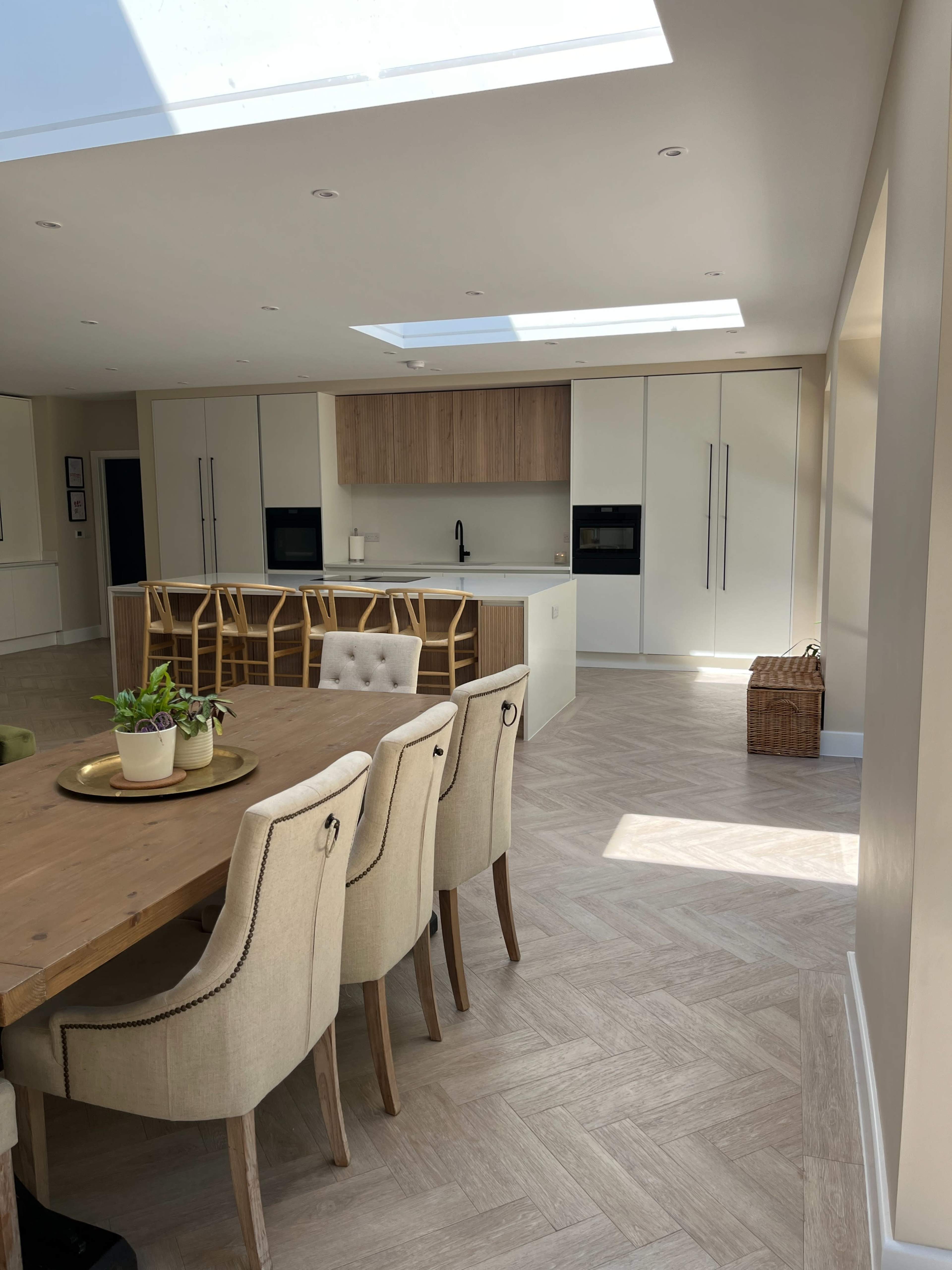 A modern kitchen with a dining area features a wooden table and beige upholstered chairs, and is complemented by light cabinetry and natural light coming through skylights.