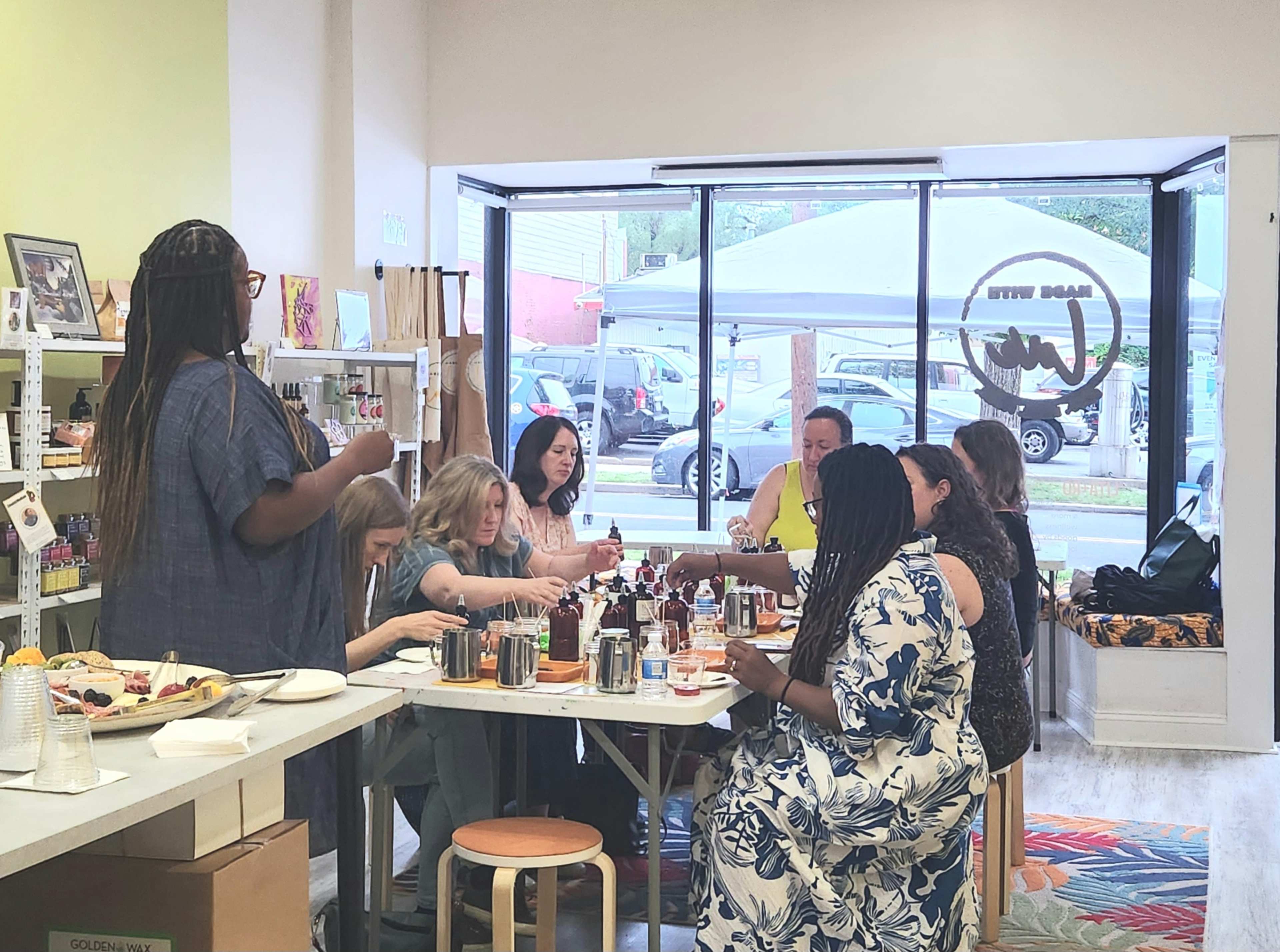 A group of people is gathered around a table, engaged in a crafting session, with various materials and beverages present.