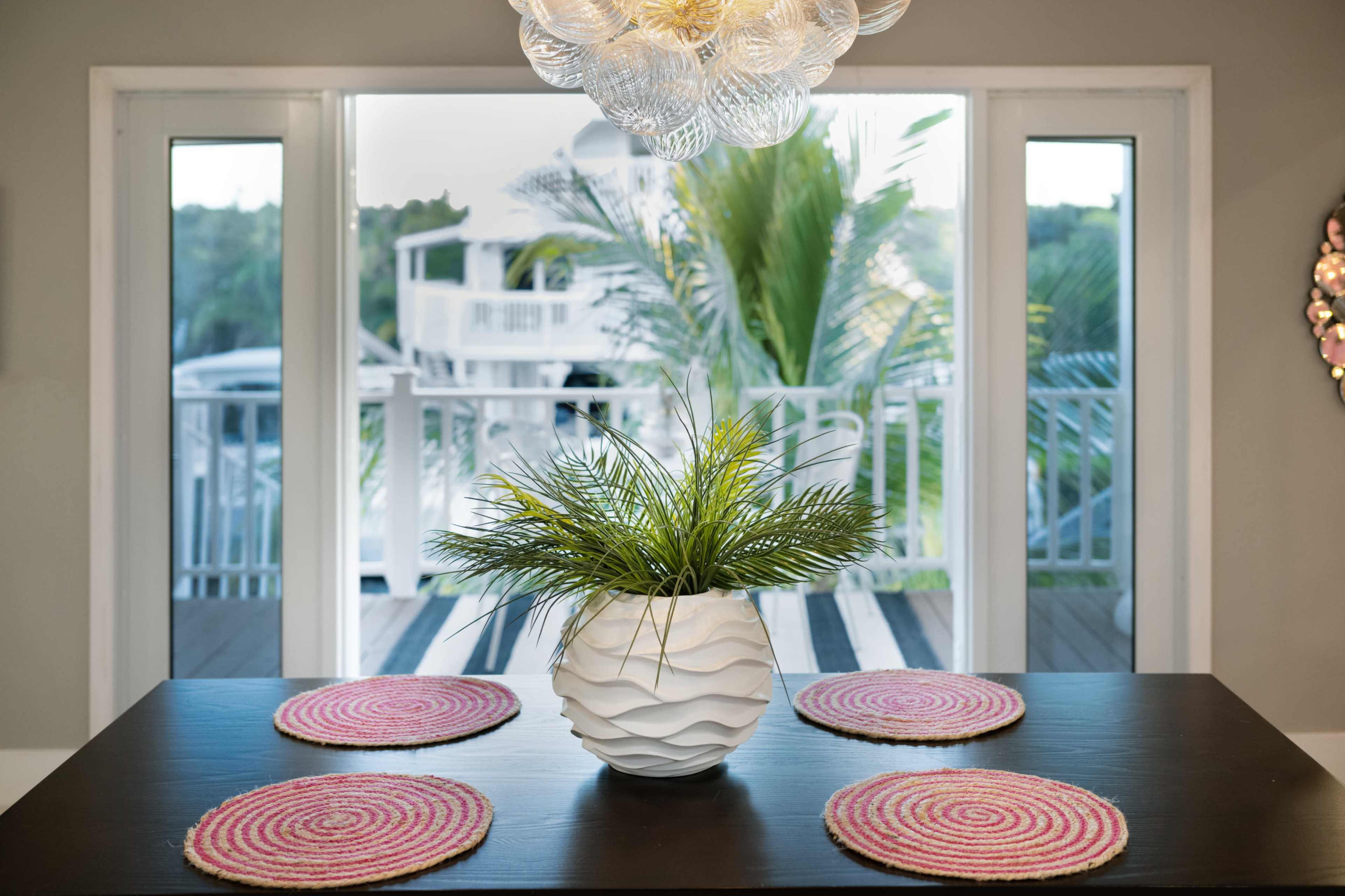 A dining table with circular pink placemats is set in front of large windows overlooking a deck and palm trees.