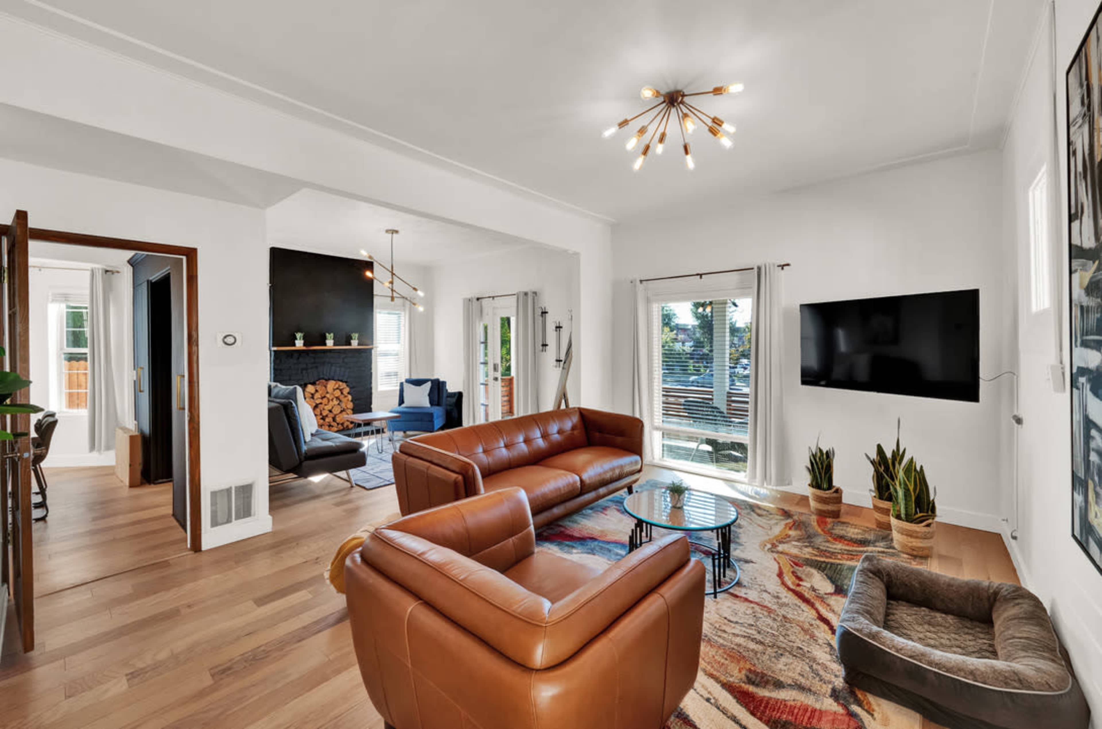 A cozy living room features brown leather sofas, a circular coffee table, a mounted television, and large windows that provide natural light.