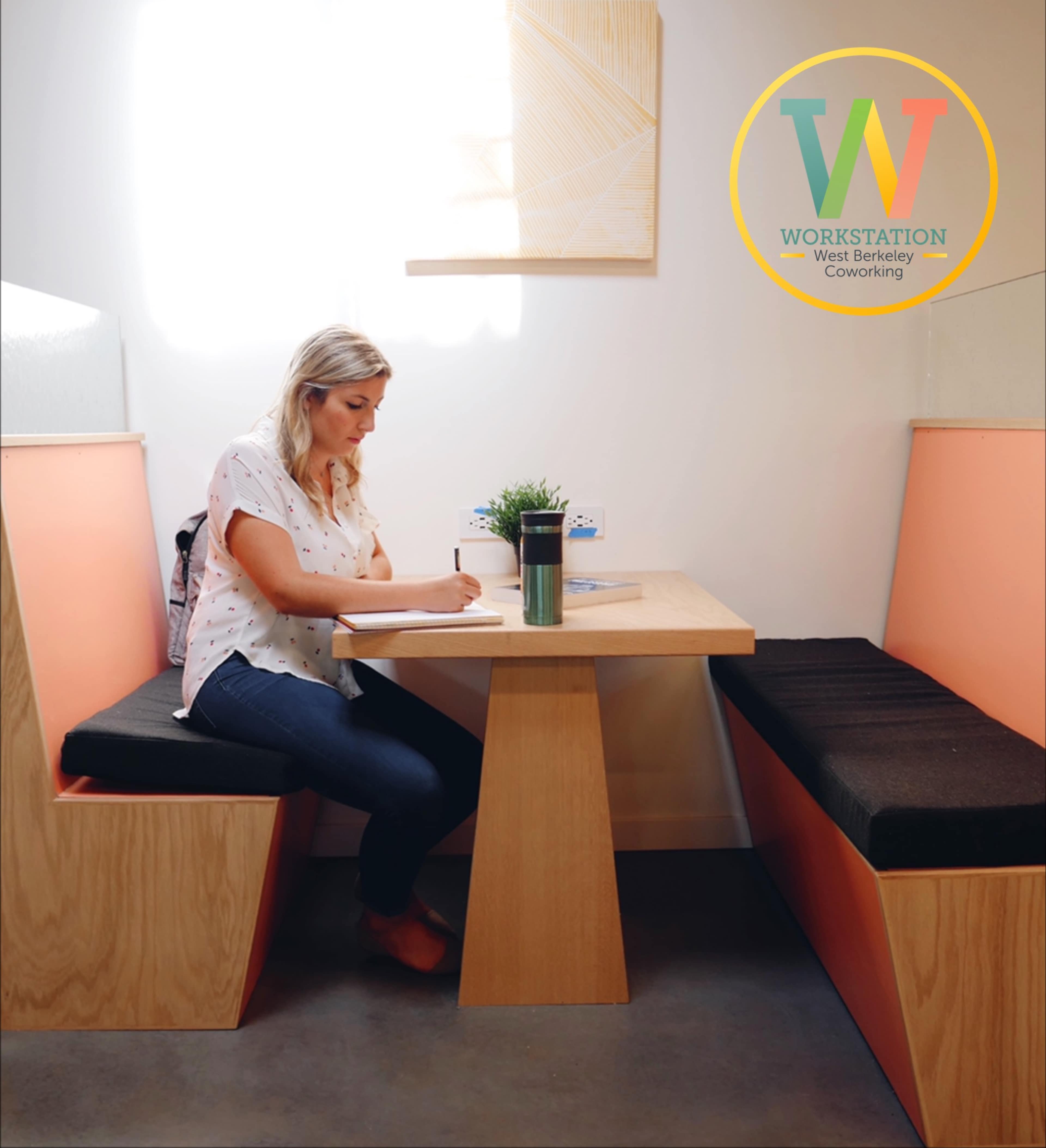A woman sits at a wooden table in a modern coworking space, writing in a notebook while surrounded by cozy seating.