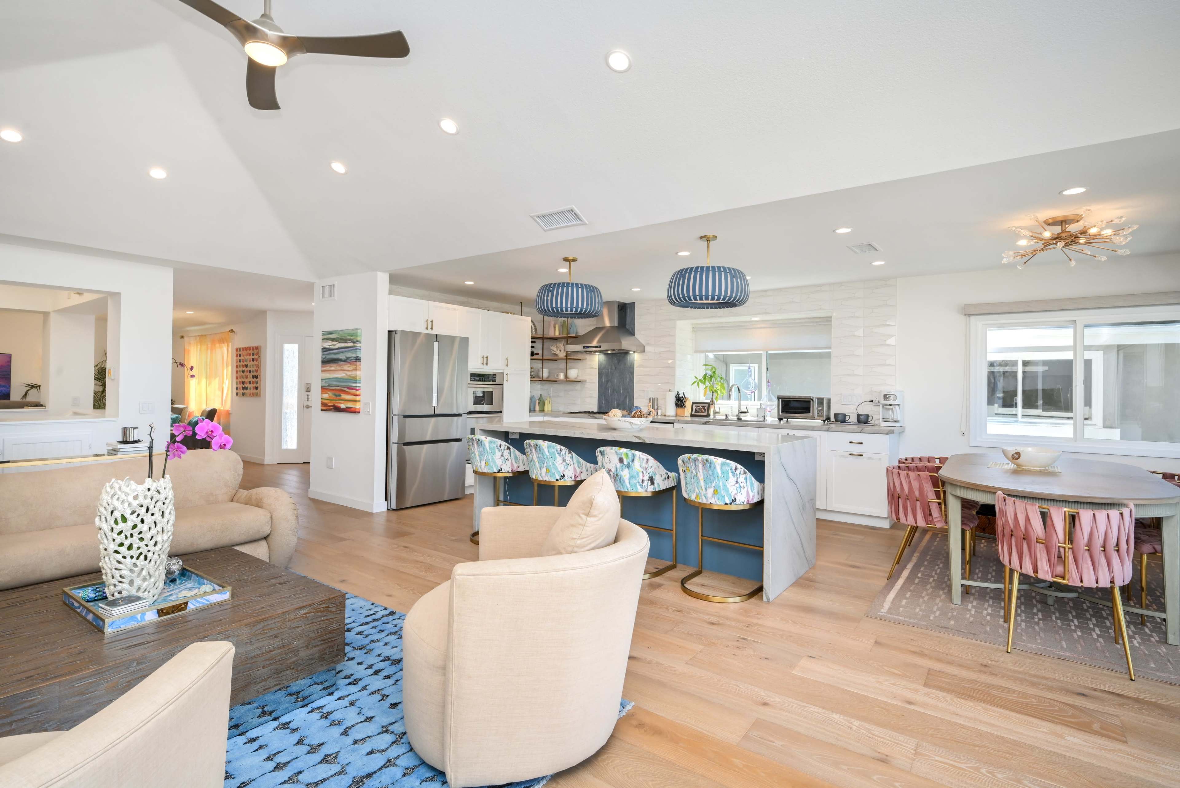 A modern open-concept kitchen and living area features sleek appliances, pastel-colored bar stools, and a dining table with pink chairs.