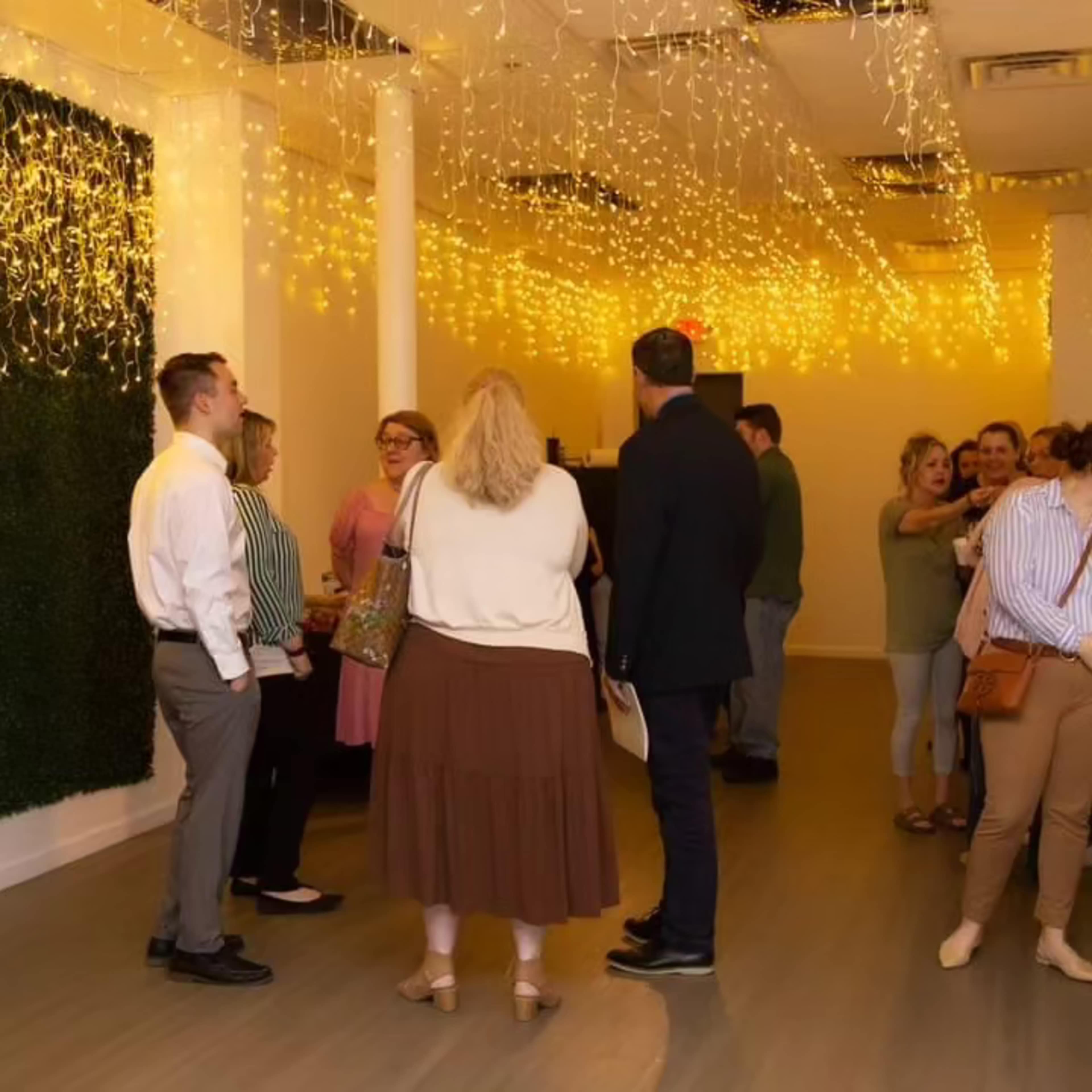 A group of people socializes in a well-lit room adorned with string lights and greenery on the walls.