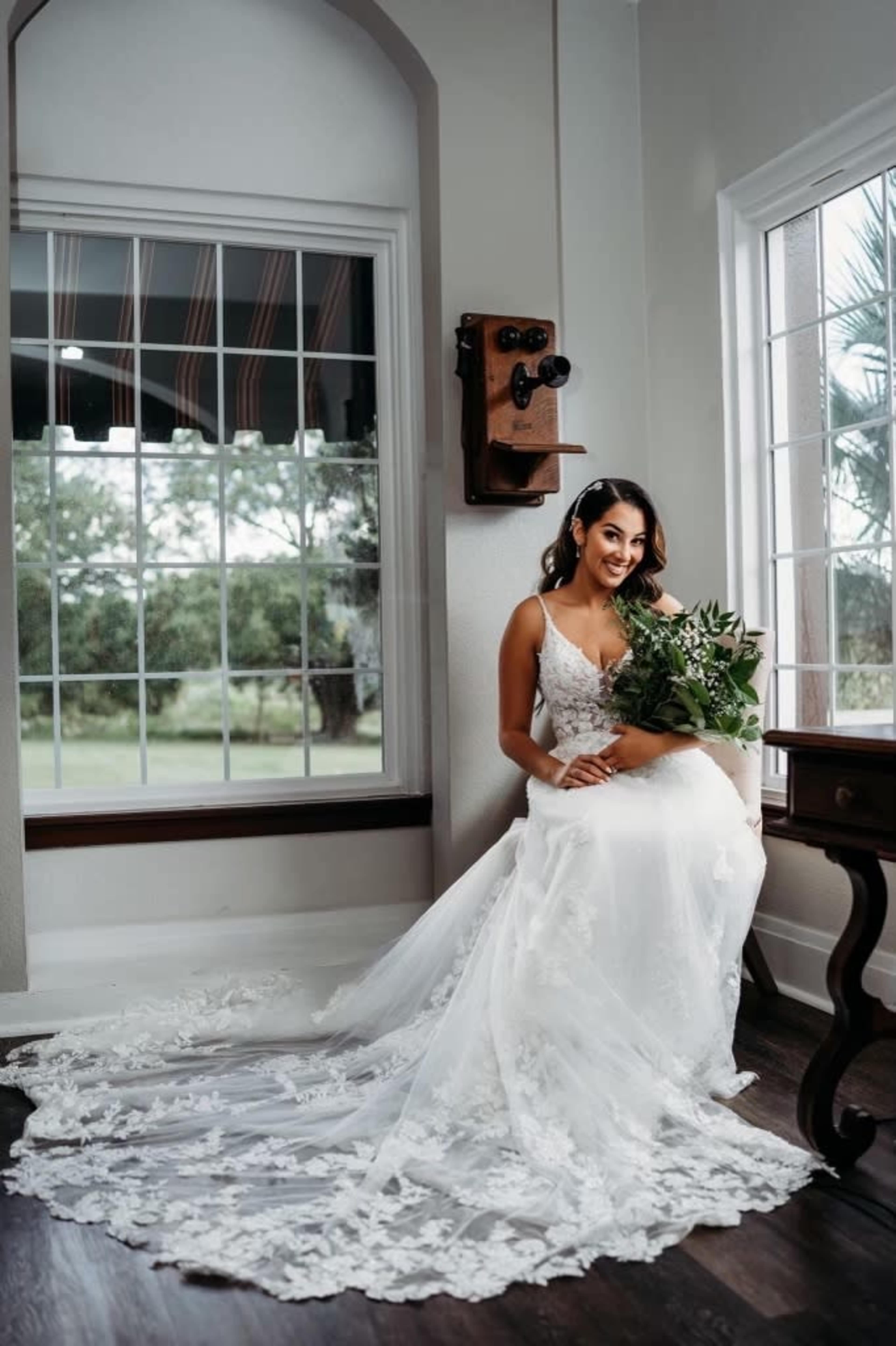 A bride in a lace gown sits elegantly on a chair near a large window, holding a bouquet of greenery.