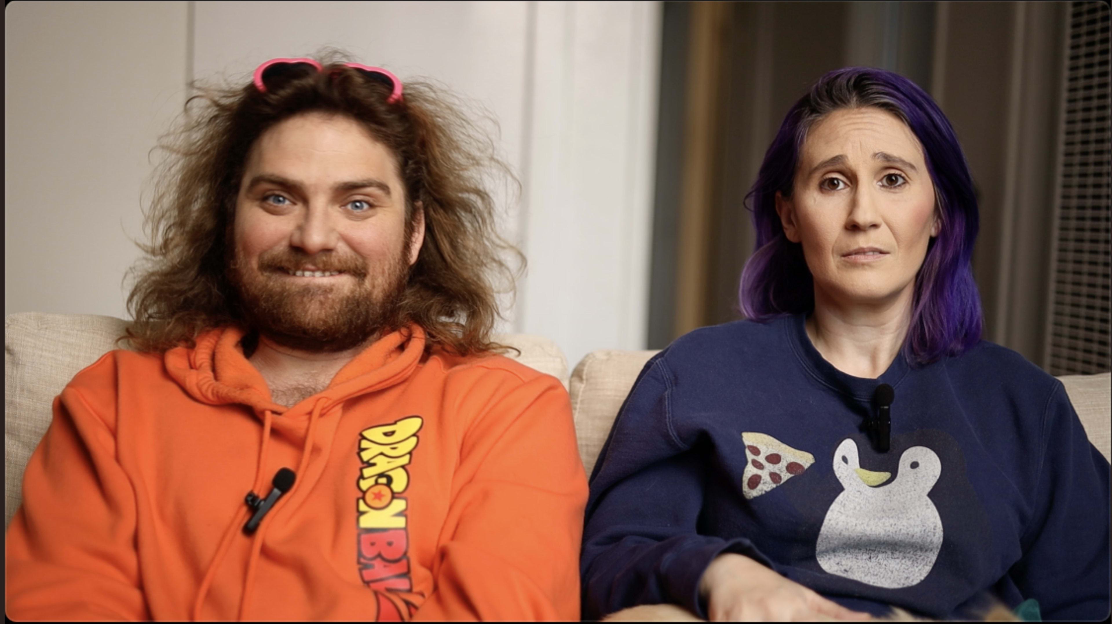 A man with curly hair wearing an orange sweatshirt and a woman with purple hair in a dark sweatshirt sit next to each other on a couch.