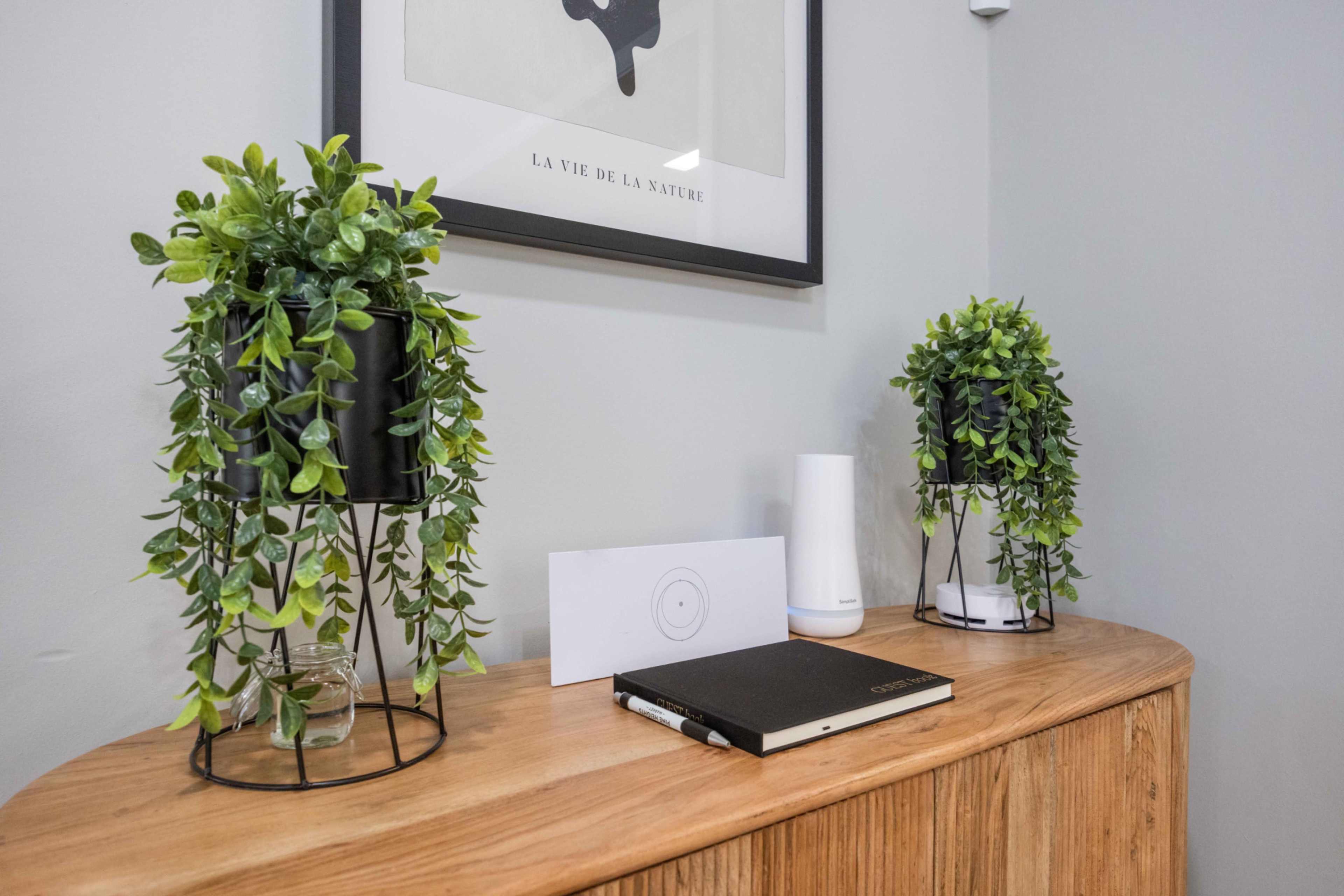 The image shows a wooden console table with two potted plants on either side, a notebook, a piece of paper, and a white smart device on top.