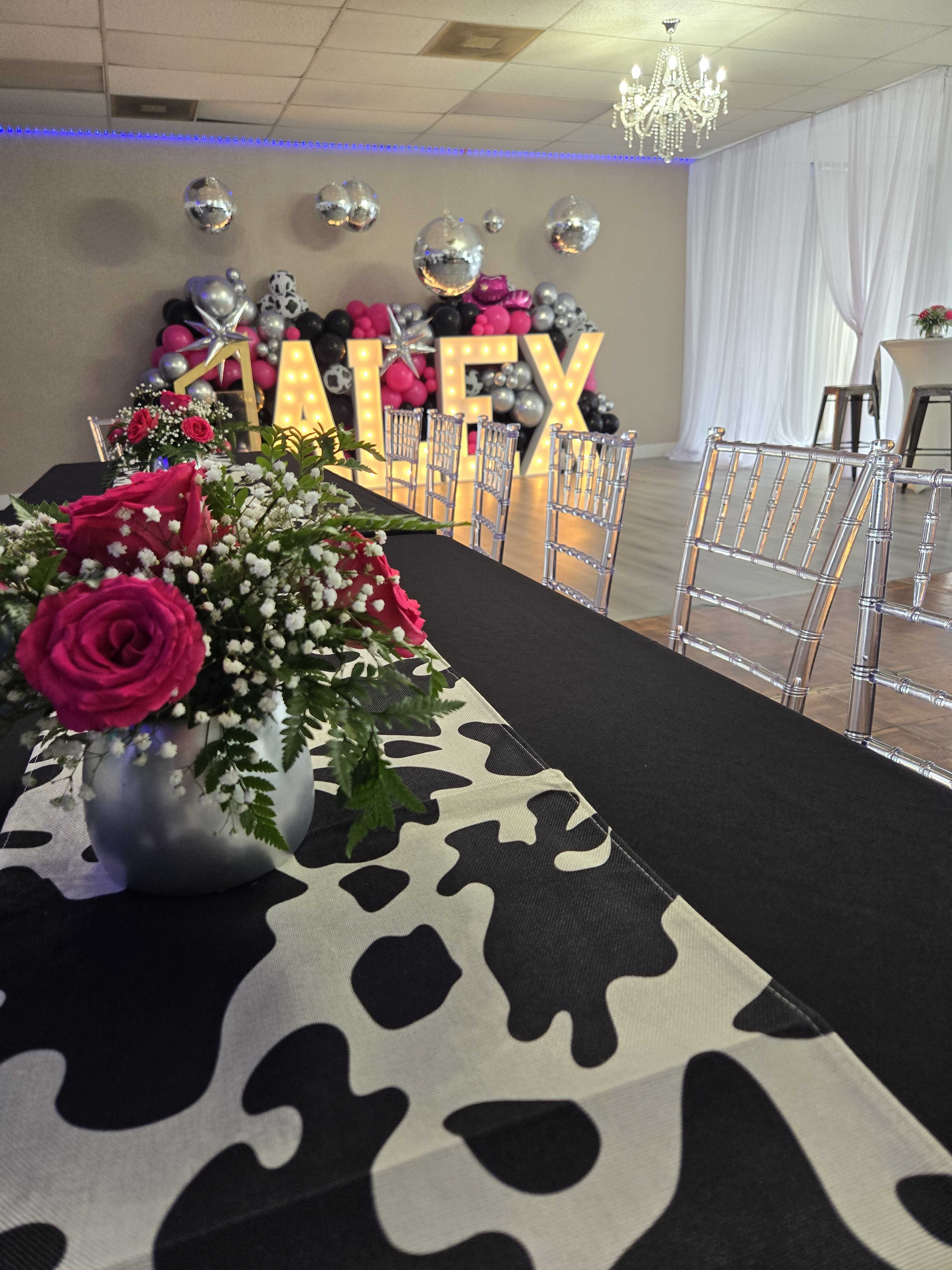 A decorated event space with a long table covered in a cow print table runner, floral arrangements, and large illuminated letters spelling "ALEX" against a backdrop of balloons and disco balls.