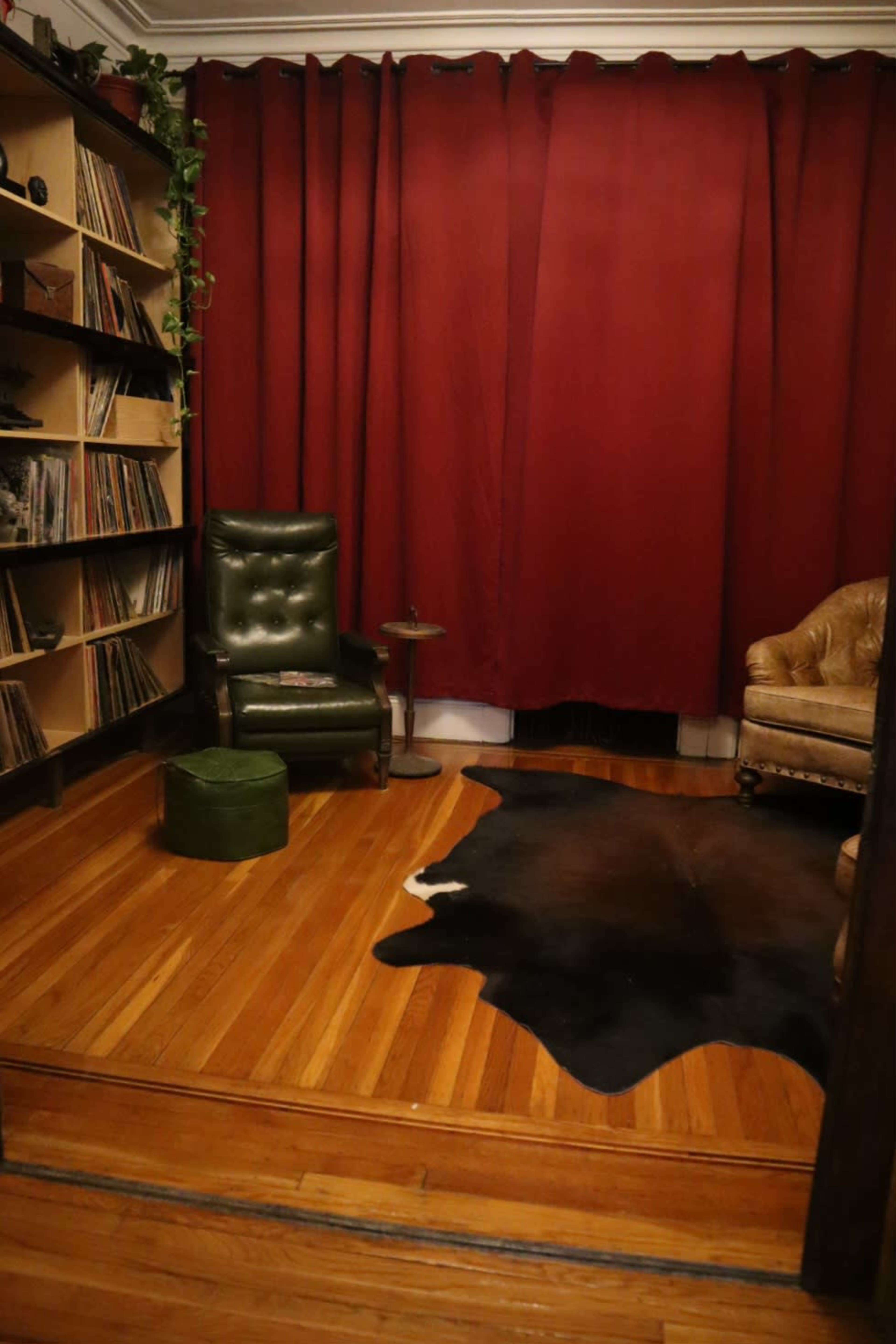 The image shows a cozy room with a vinyl record shelf, a green upholstered chair, a brown leather armchair, and a cowhide rug on wooden flooring, all set against red curtains.