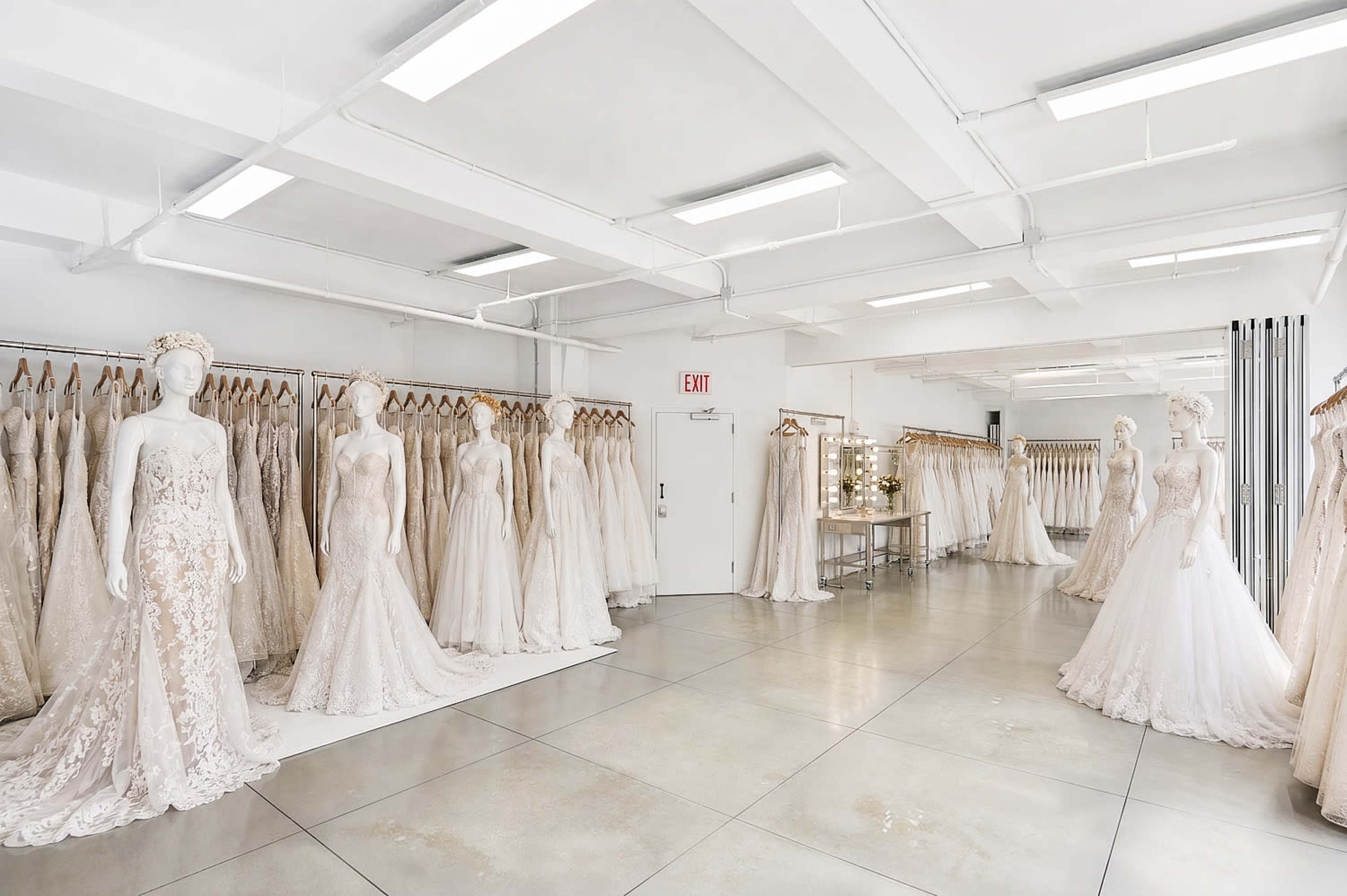 The image shows a bridal boutique with several wedding dresses displayed on mannequins and racks in a well-lit, spacious room.