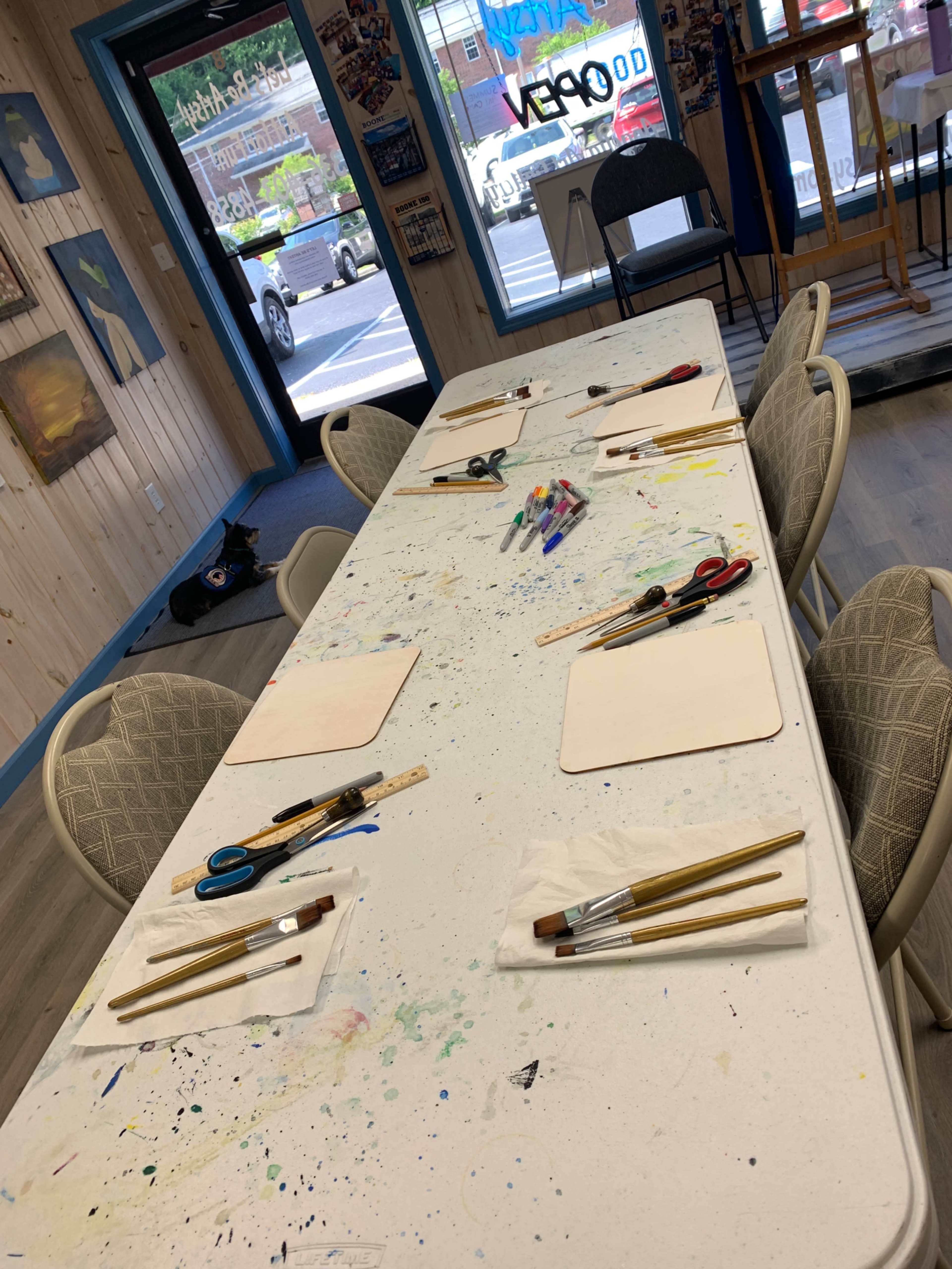 A long table is set with paintbrushes and materials, surrounded by chairs in an art studio.
