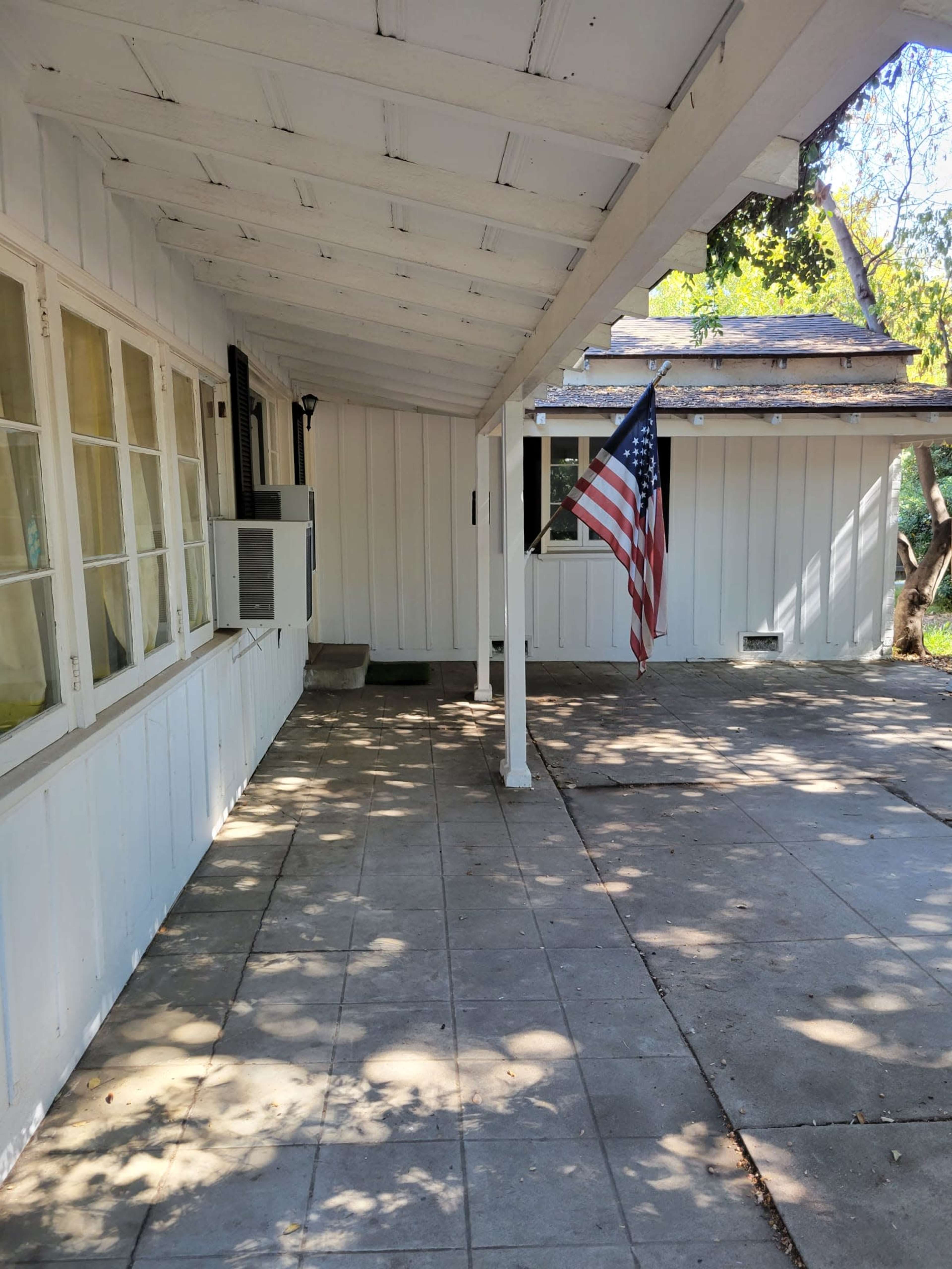 A white house exterior with a porch featuring a flagpole holding an American flag, on a sunny day with dappled shadows on the concrete floor.