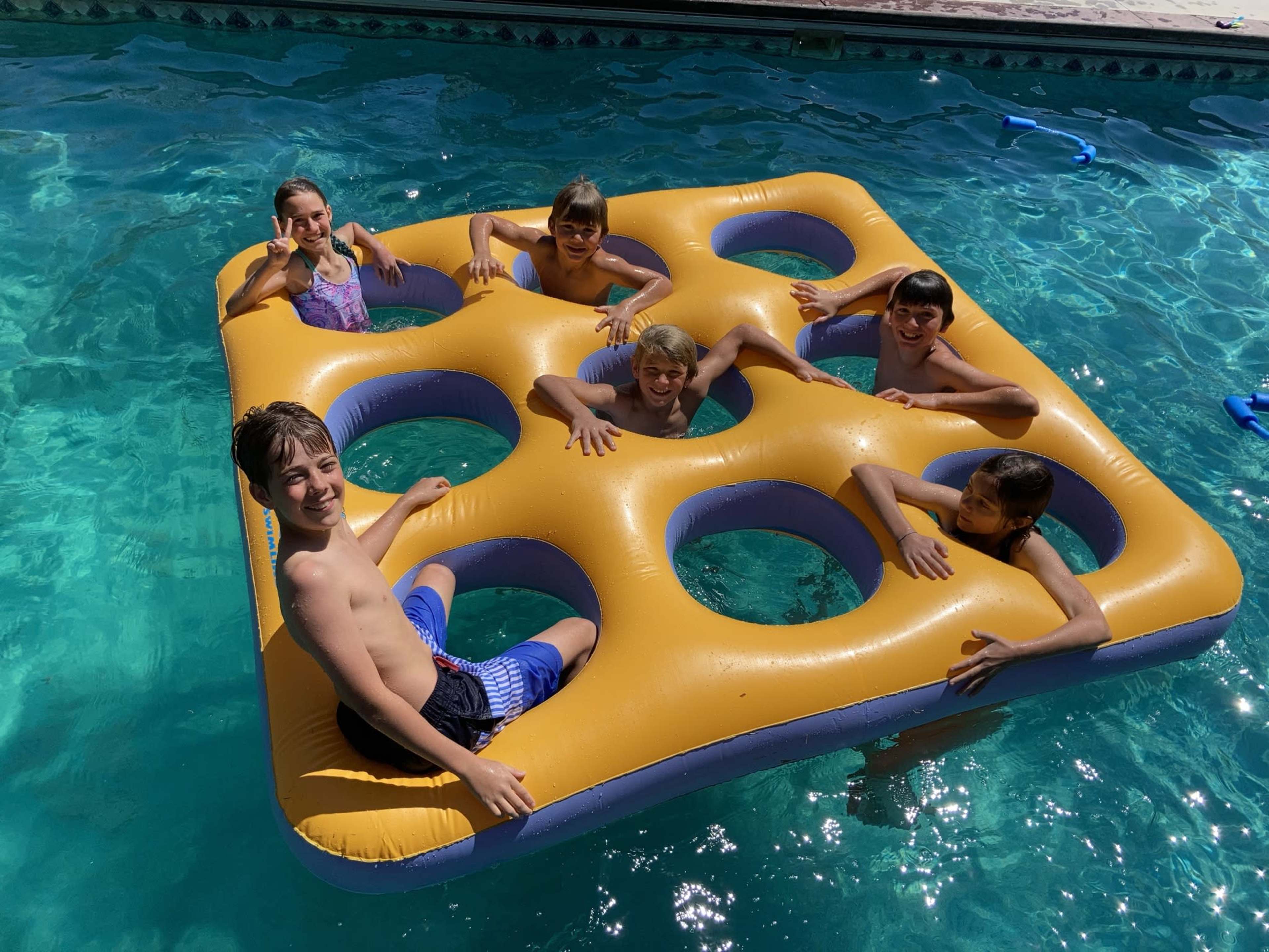 A group of six children is playing on a large, yellow square float with holes in a swimming pool.
