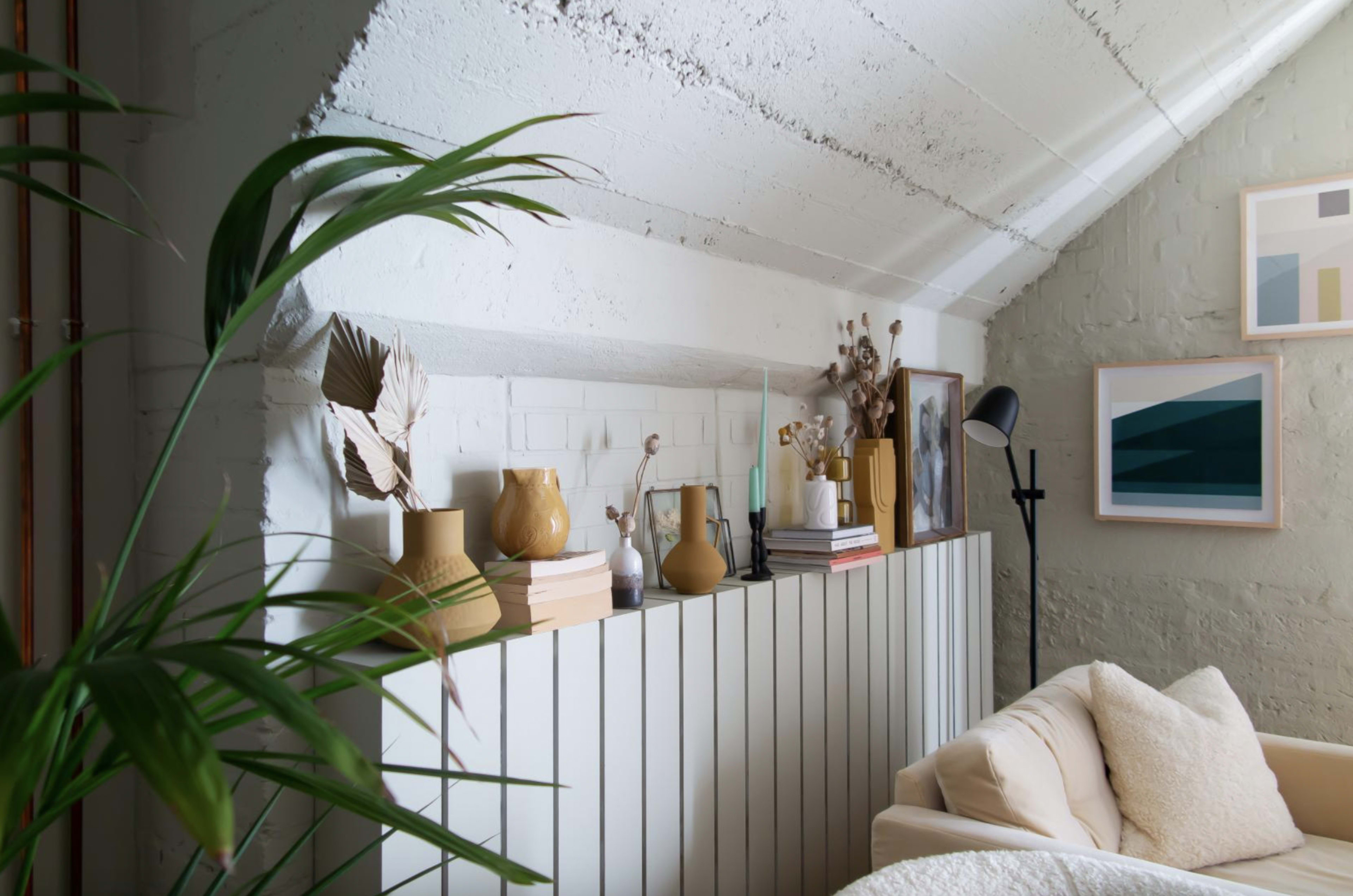 A cozy living space with a low shelf displaying decorative items and books, alongside a tall green plant and framed artwork on the wall.