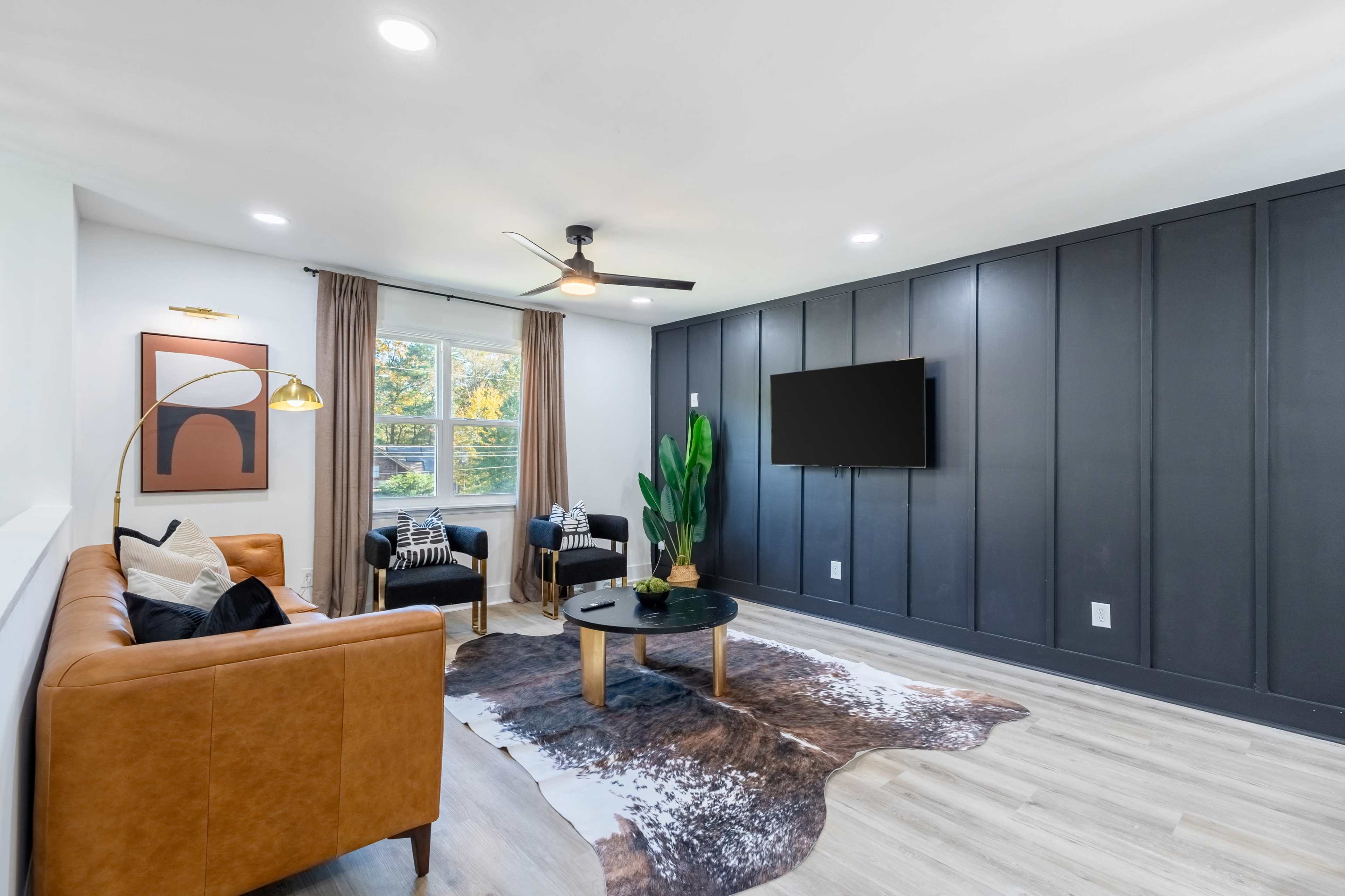 The image shows a modern living room with a brown leather sofa, two black armchairs, a wooden coffee table, and a television mounted on a dark paneled wall.