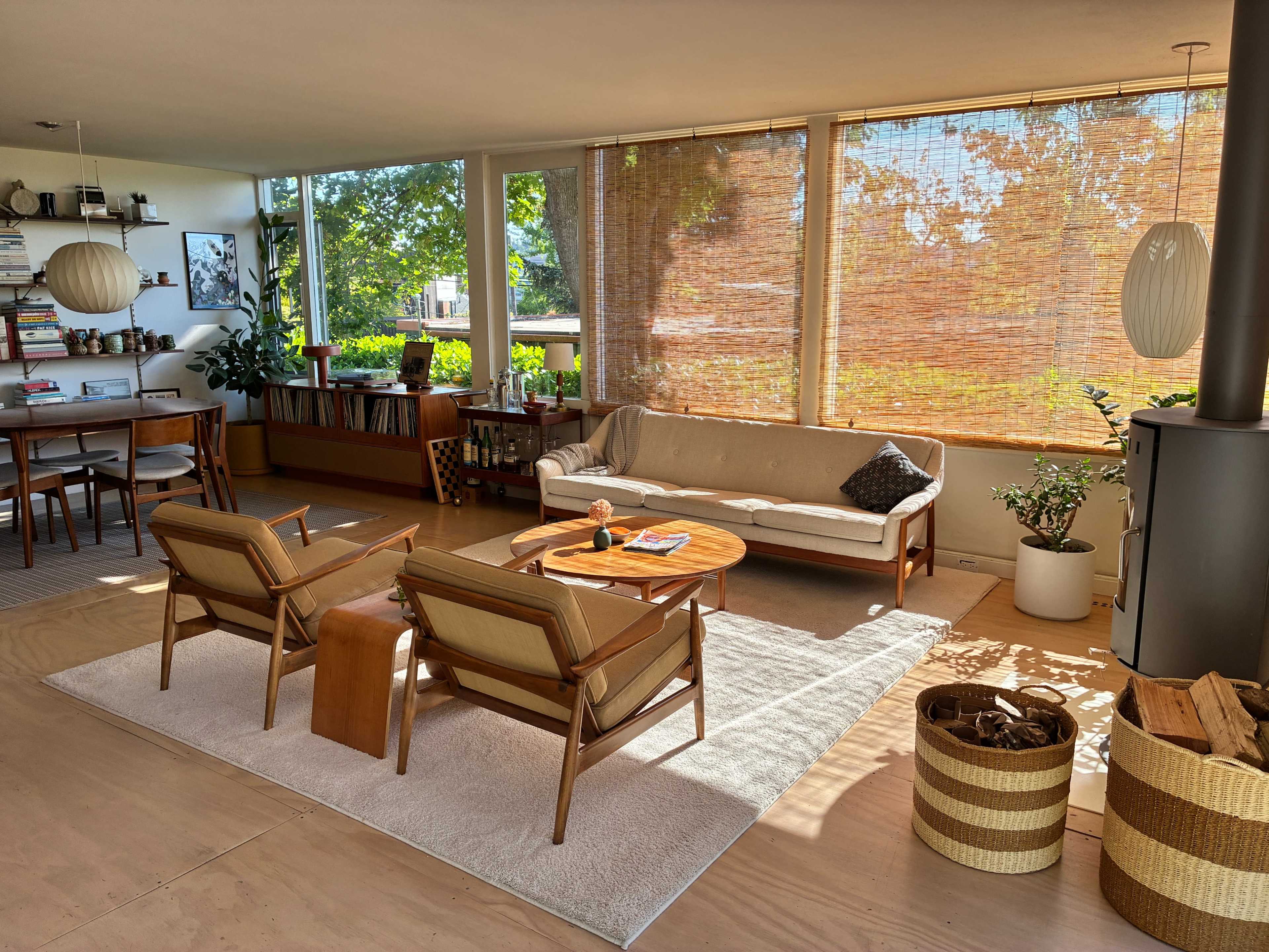 A spacious living room features a sofa, two chairs, a round coffee table, and large windows with natural light, along with decorative plants and a dining area in the background.