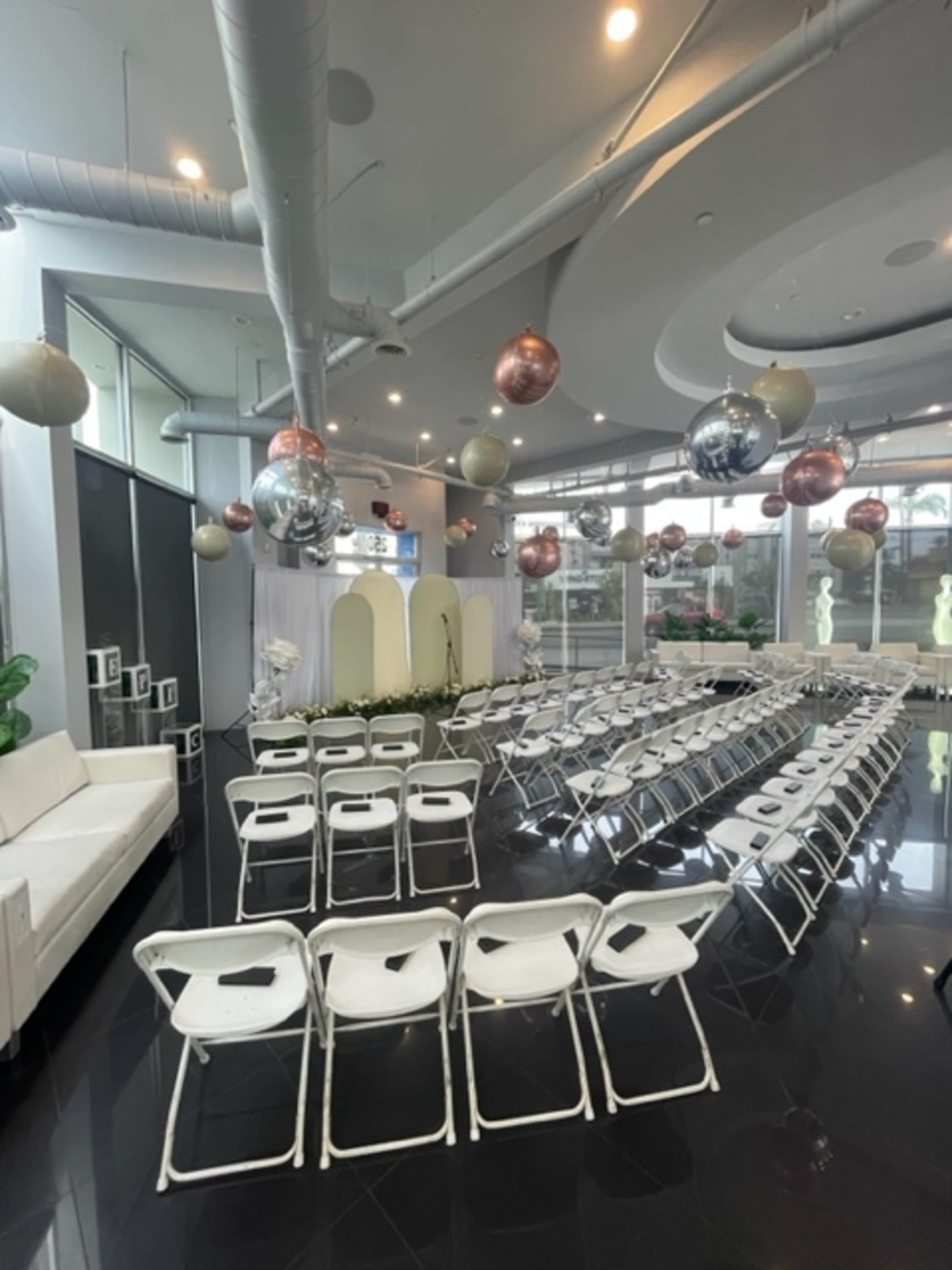 The image shows a modern event space arranged with white chairs facing a decorated stage, complemented by hanging decorative orbs.