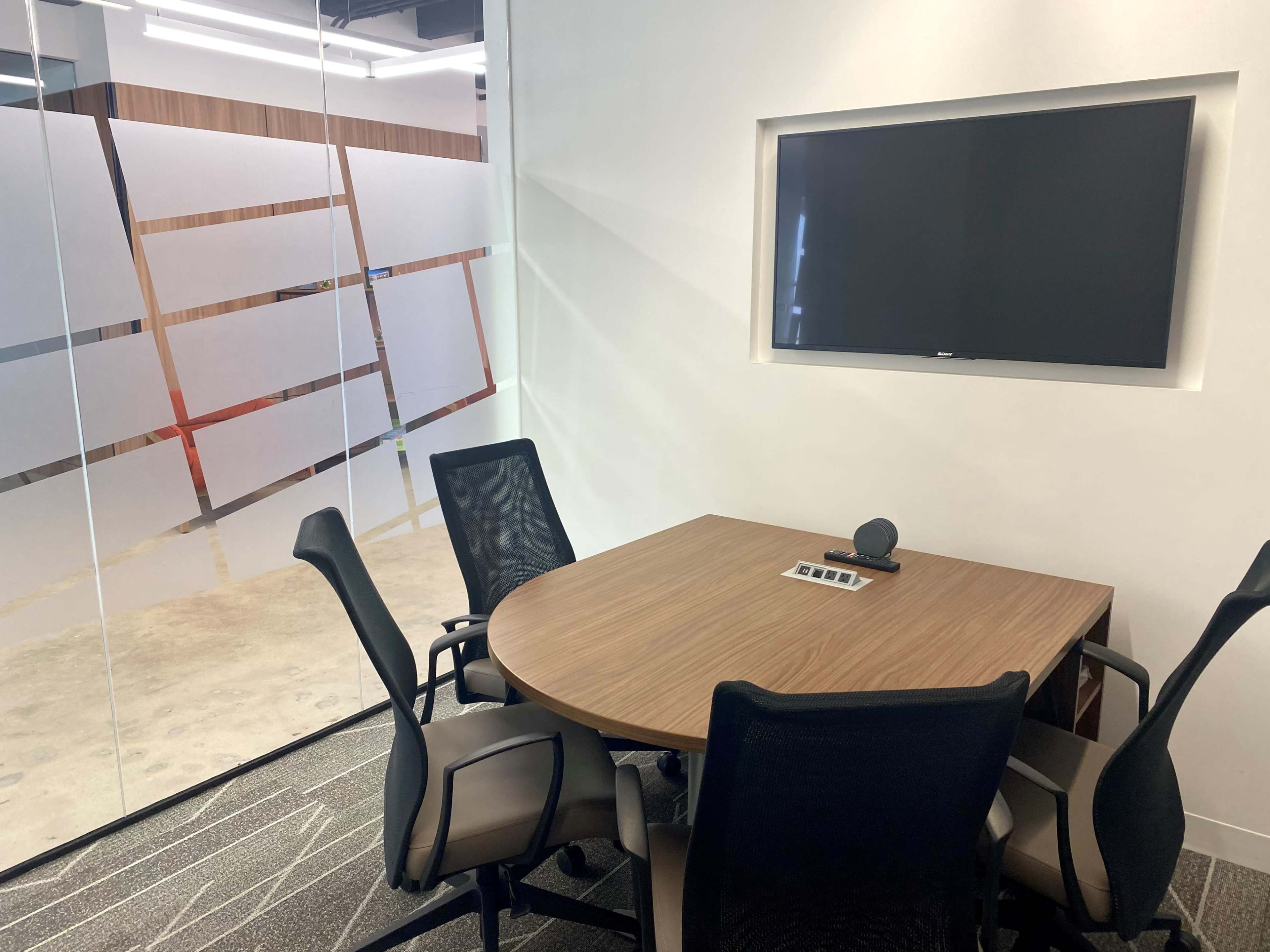 A small conference room features a round wooden table, five chairs, a wall-mounted television, and a glass panel with geometric designs.