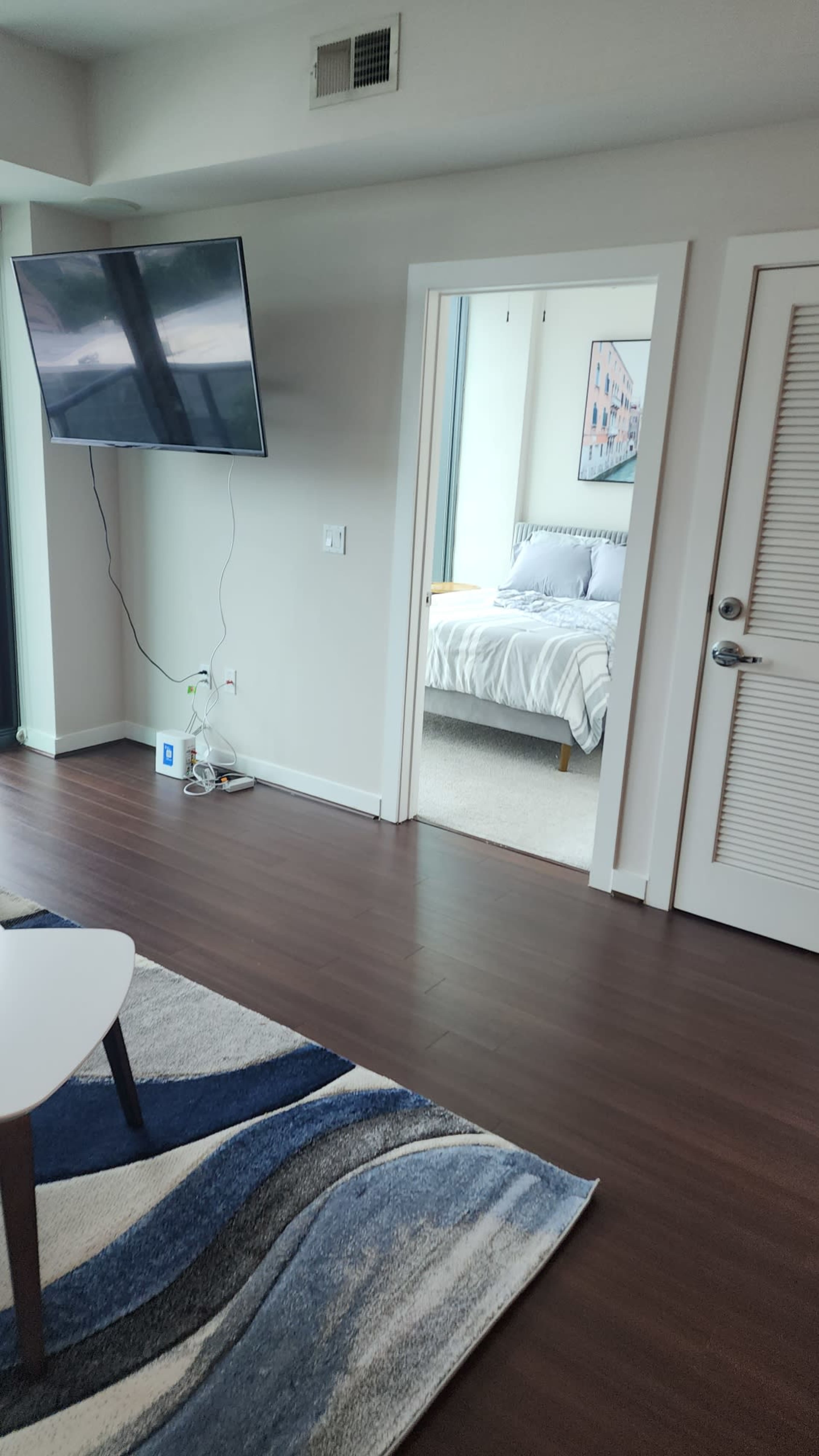 A modern living area with a wall-mounted TV, a doorway leading to a bedroom, and a patterned rug on a hardwood floor.