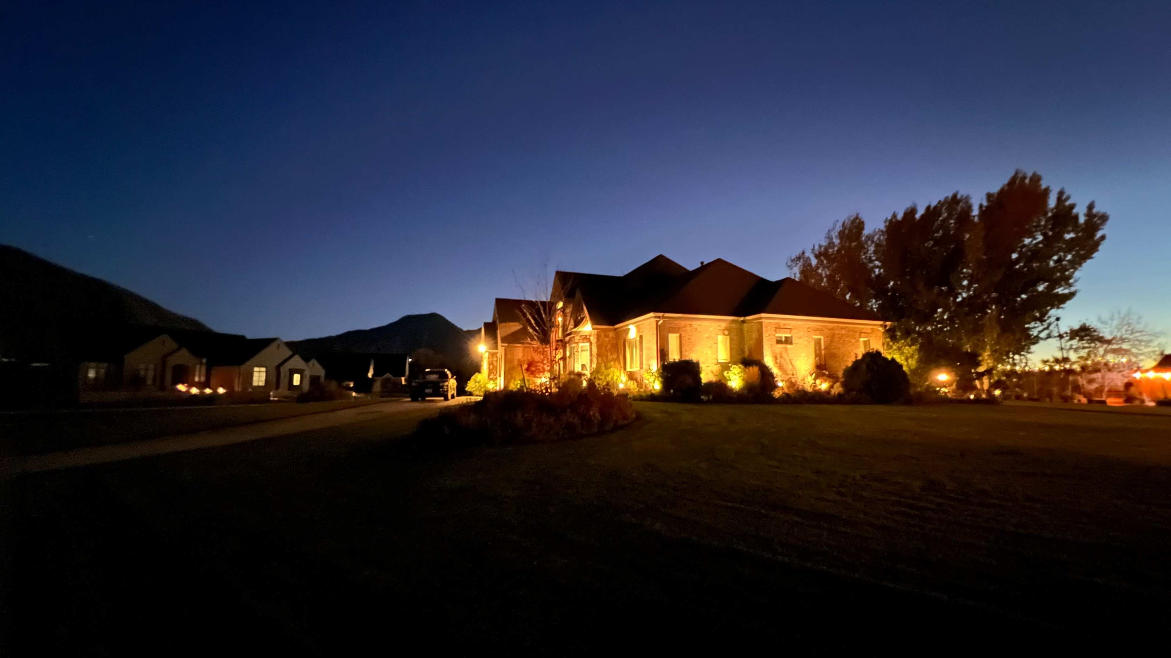 A well-lit house sits in a rural setting at dusk, with mountains in the background and landscaping illuminated by warm lights.