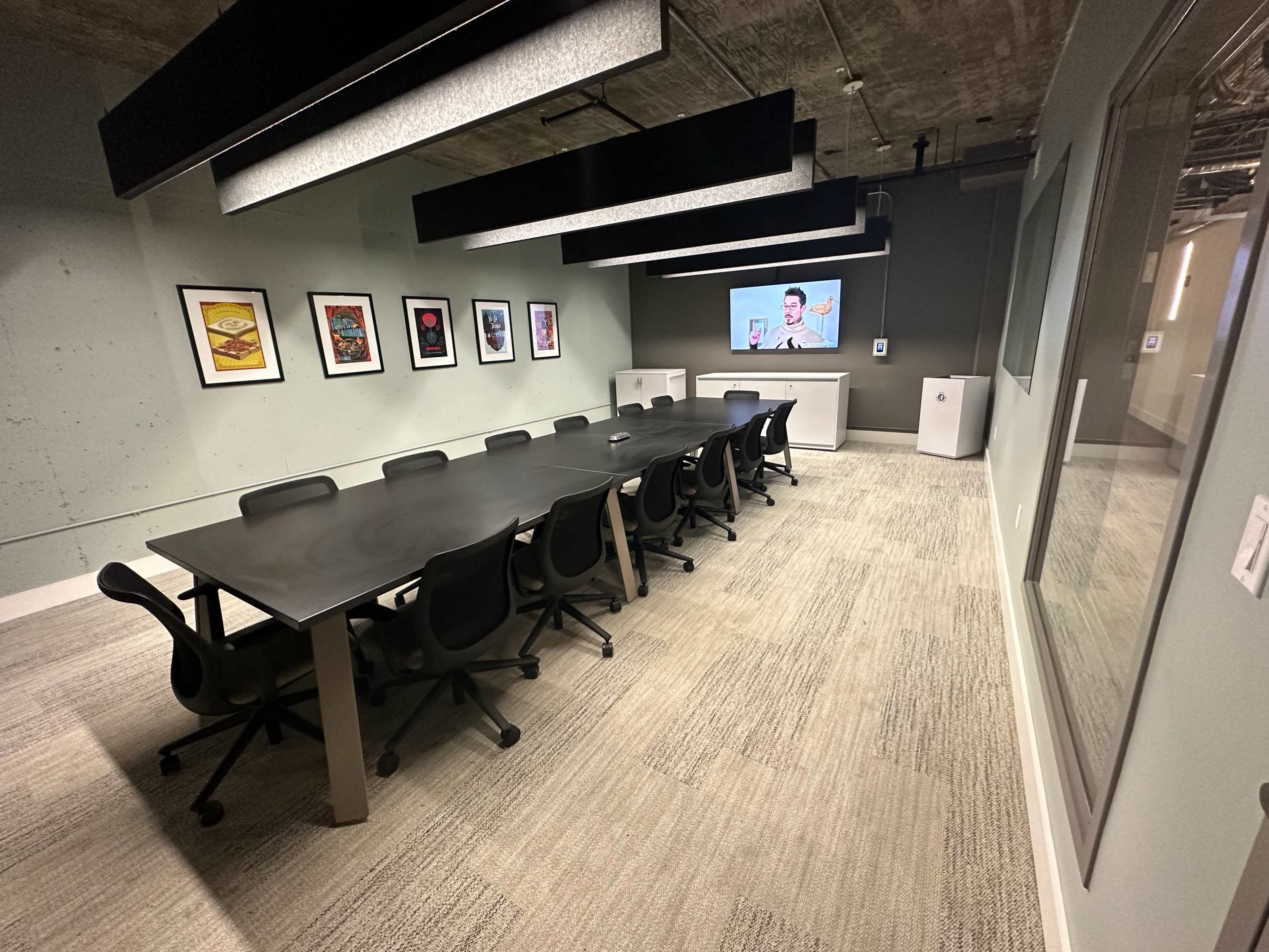 A long rectangular conference table is set up in a modern meeting room with surround black chairs, artwork on the walls, and a presentation screen at one end.