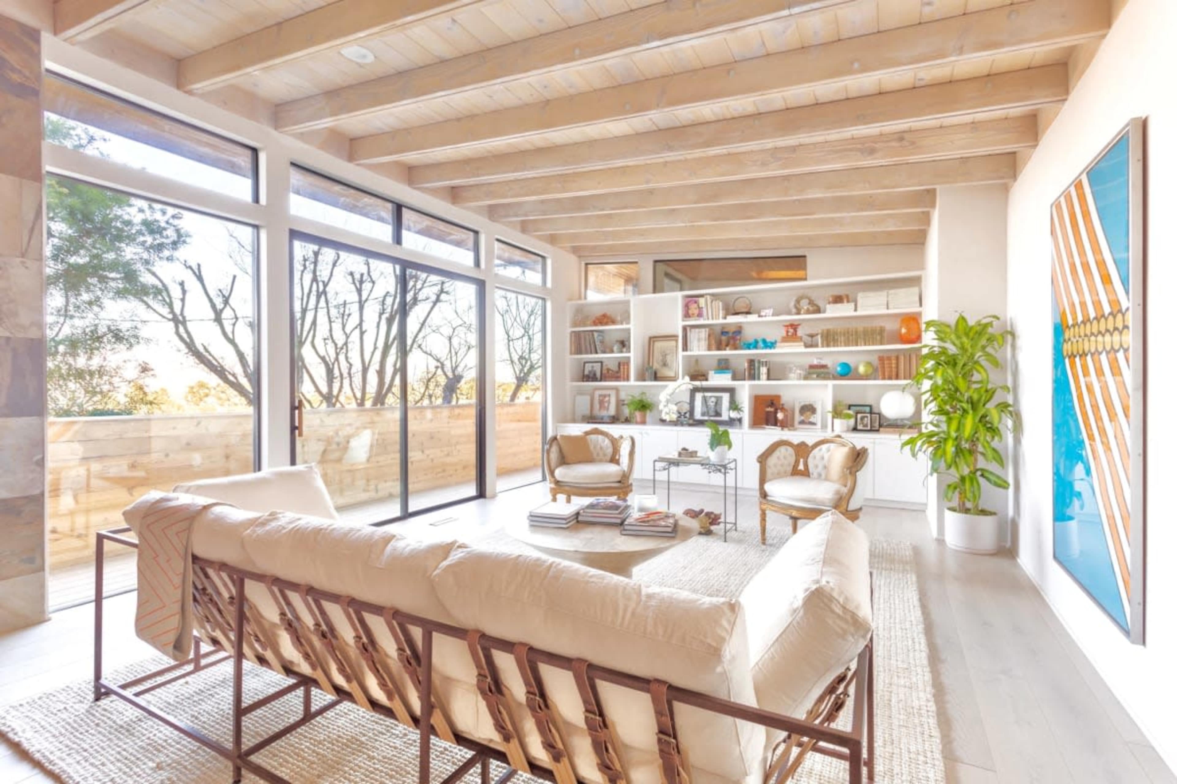 The image shows a modern living room with a light wooden ceiling, large glass doors, and shelves filled with books and decorations.