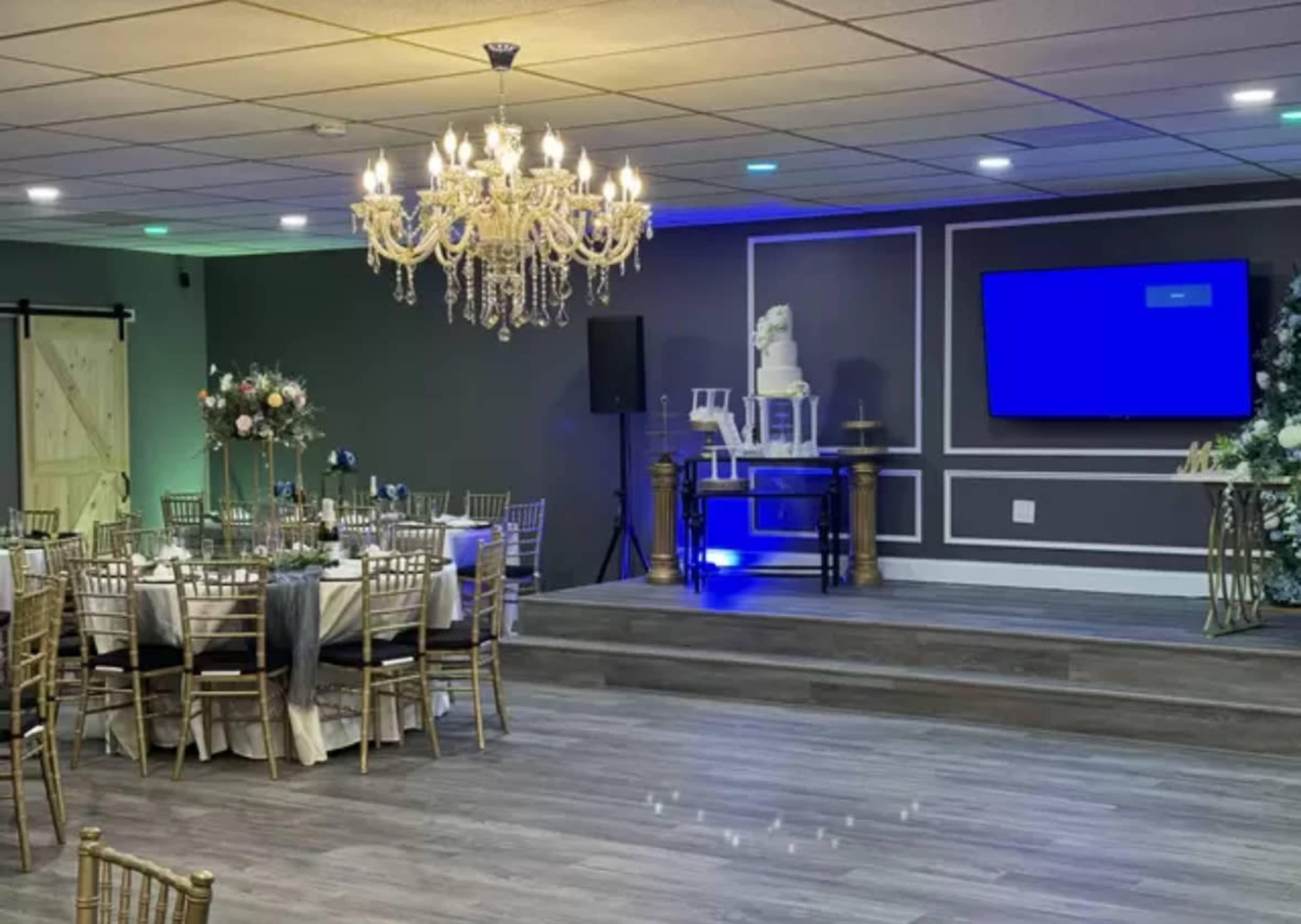 The image shows a decorated event space with round tables set for dining, a stage featuring a cake display, and a large screen in the background.