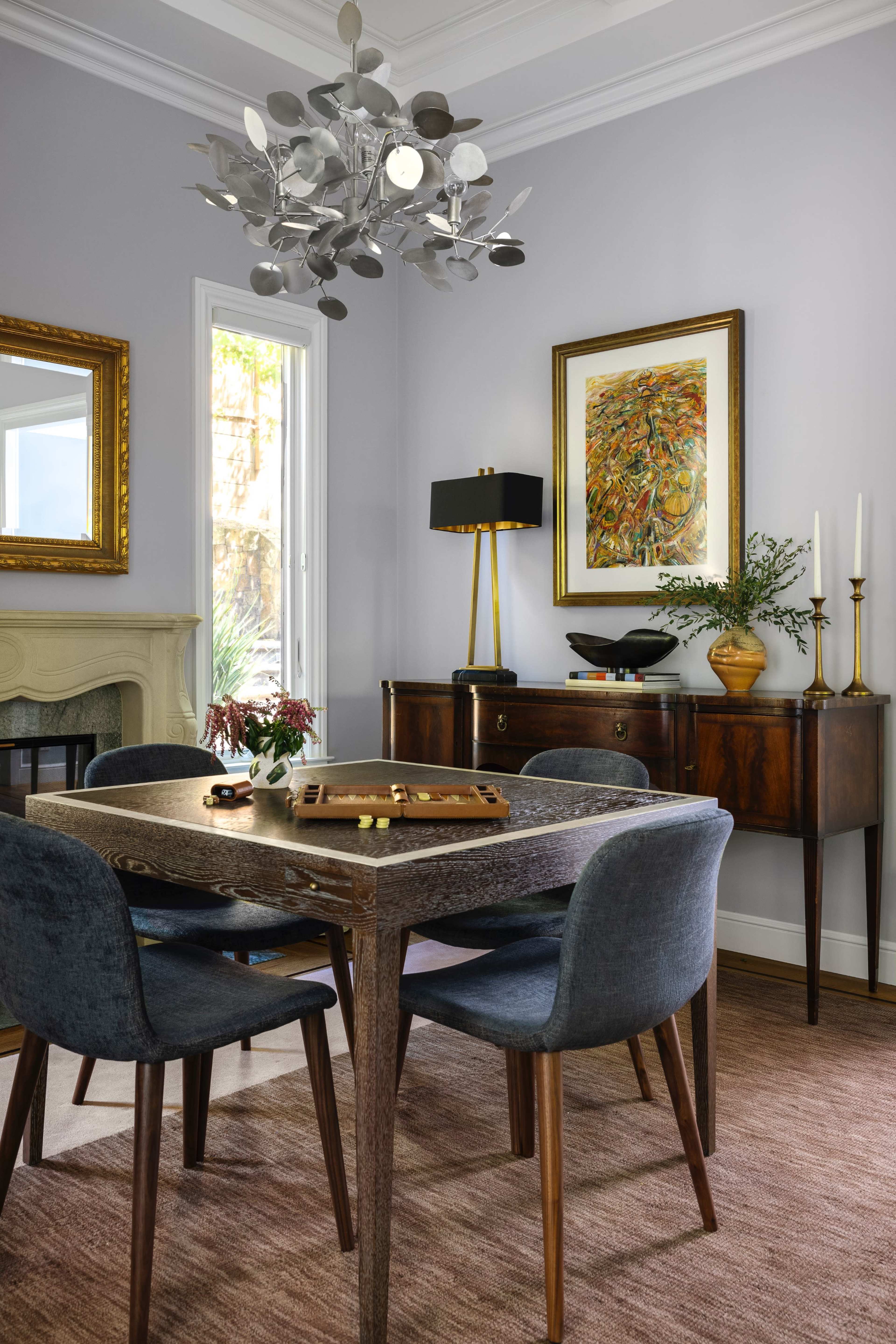 A dining area features a wooden table surrounded by blue upholstered chairs, a modern chandelier overhead, a decorative painting on the wall, and a sideboard with a lamp and plant.