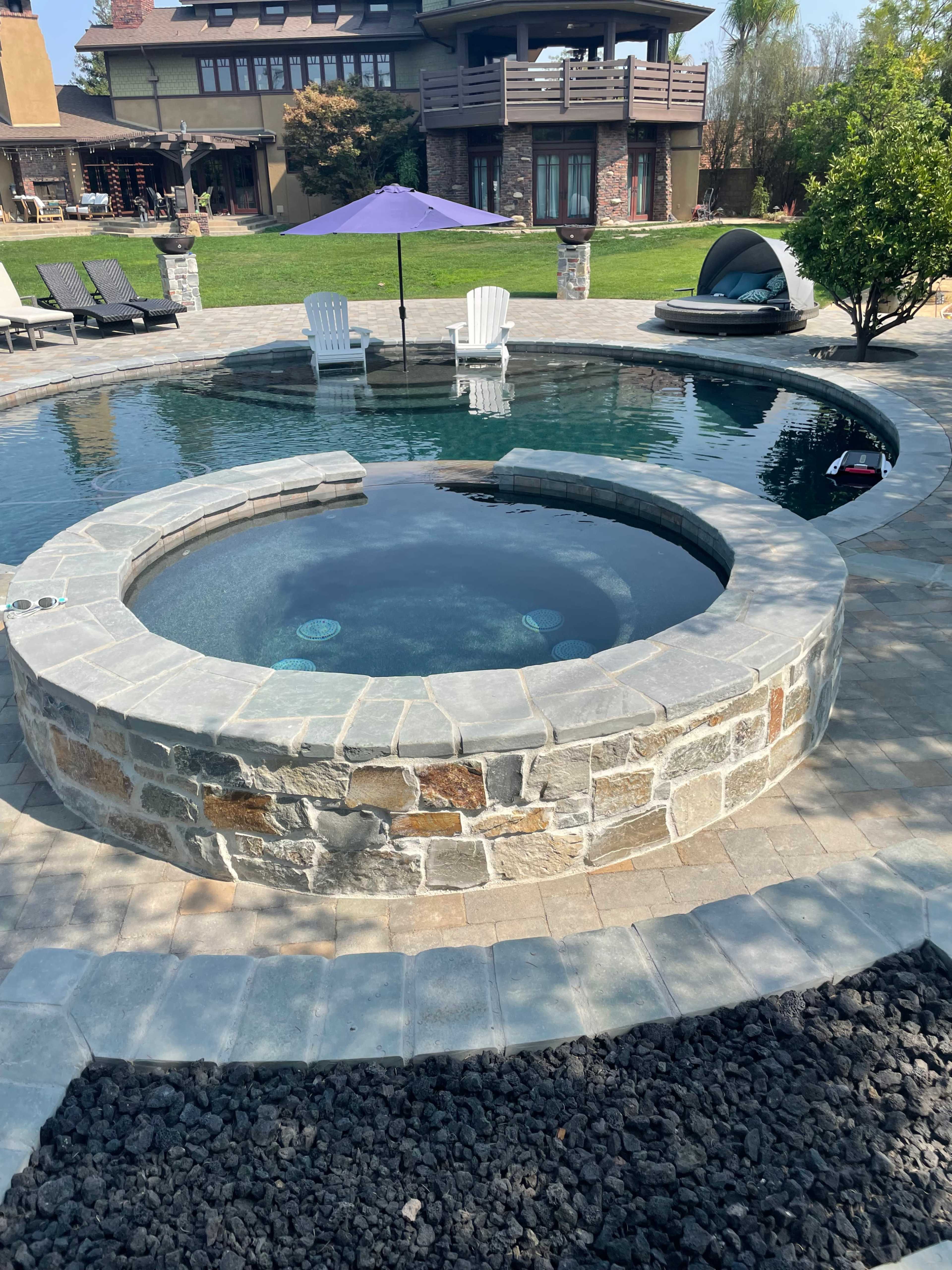 The image shows a circular stone hot tub situated next to a rectangular swimming pool in a sunny backyard with lounge chairs and a purple umbrella.