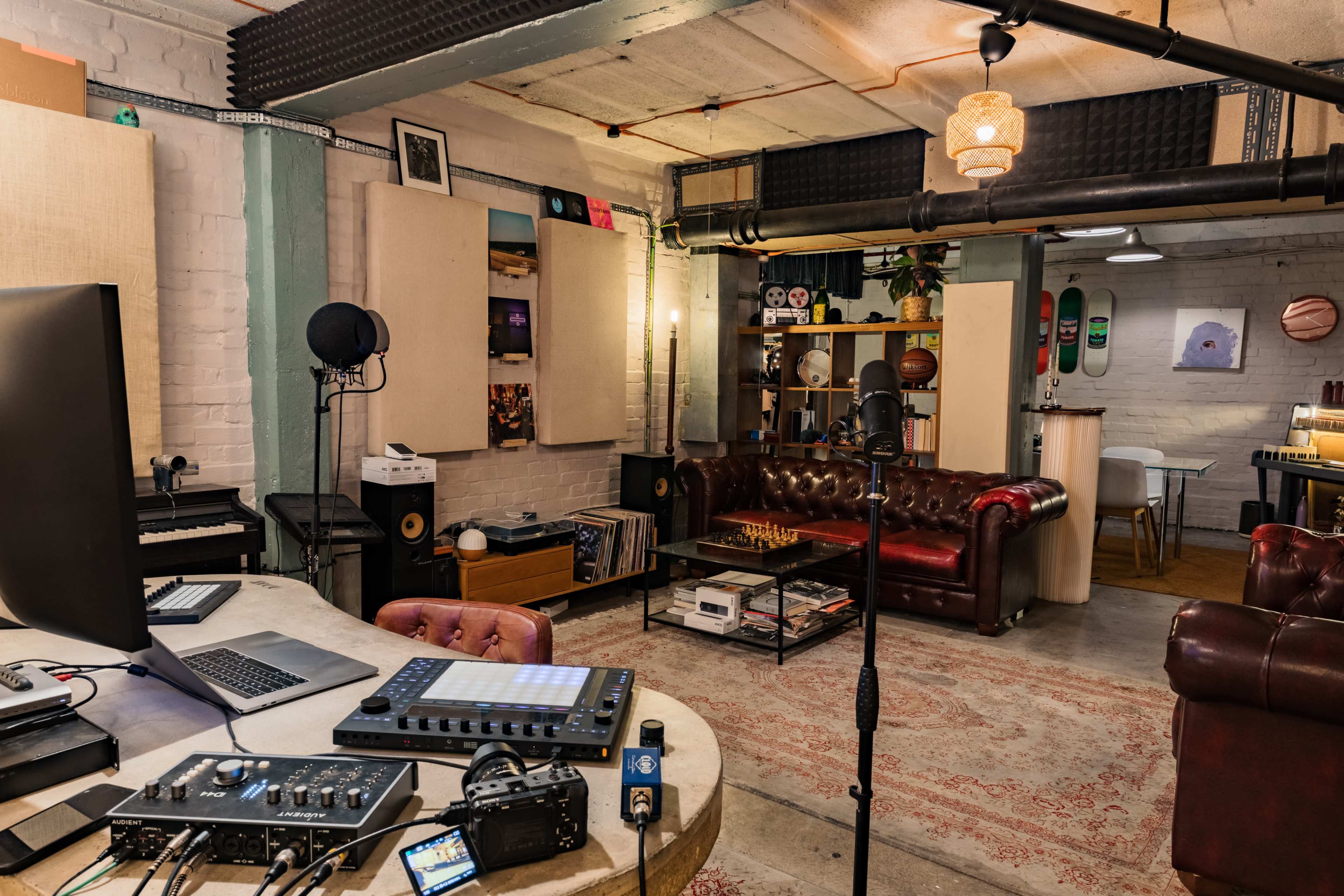 The image shows a music recording studio with a mix of electronic equipment, a microphone, and comfortable seating, surrounded by shelves and artistic decor.