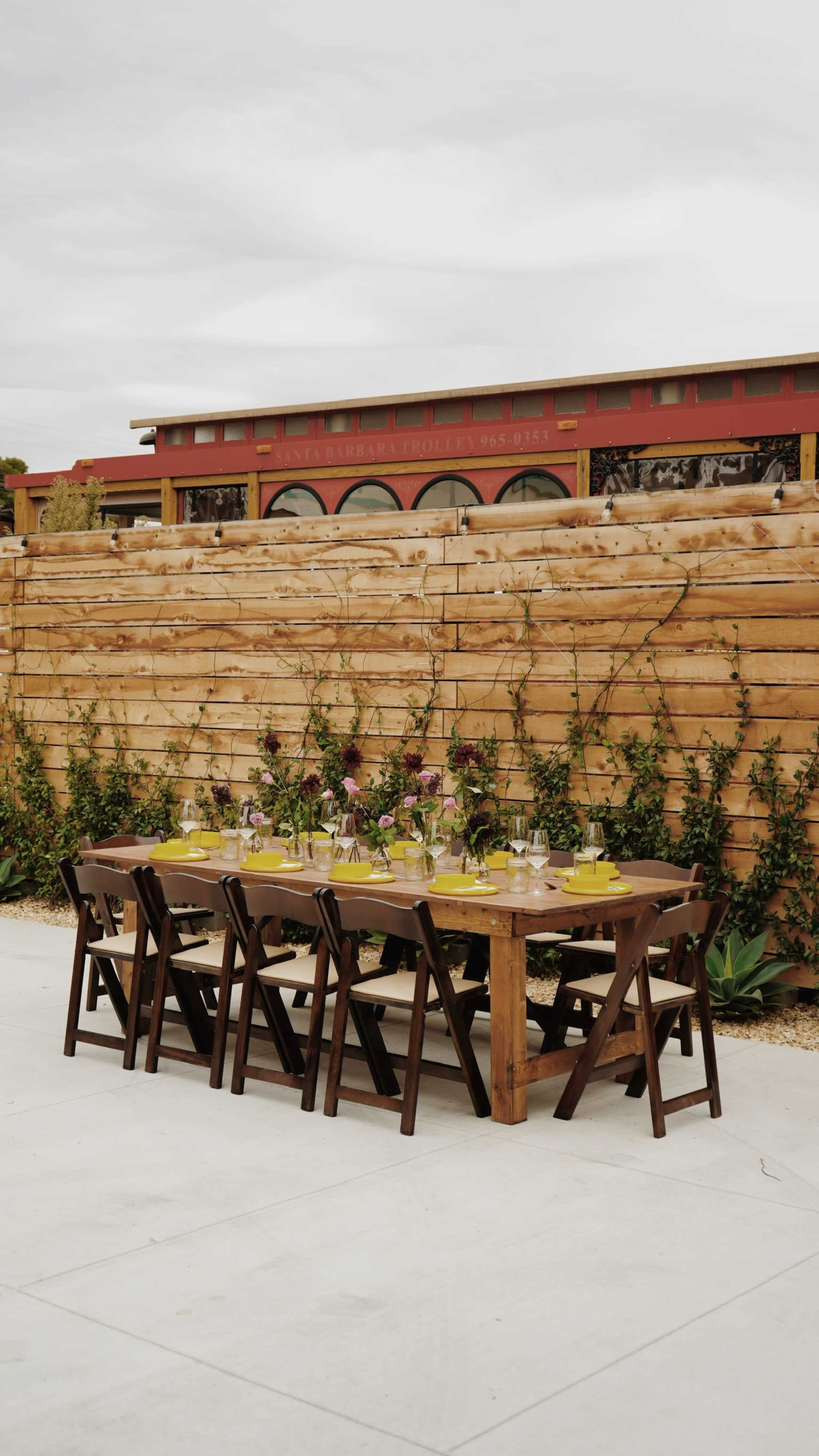 A long wooden dining table is set with decorative plates and flowers against a backdrop of a wooden fence and a red building.