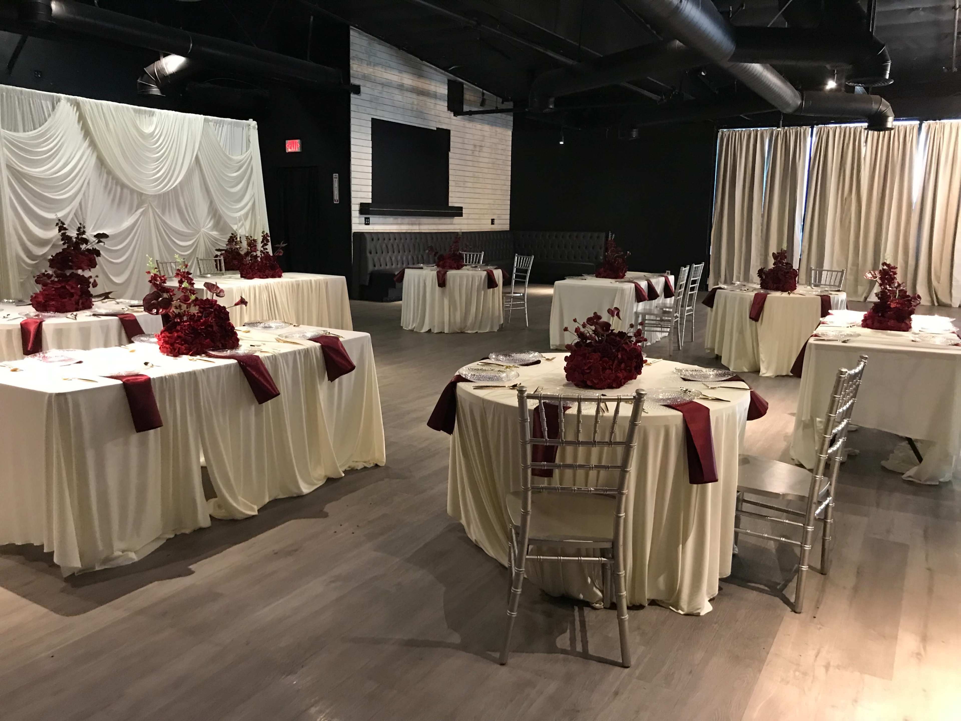 The image shows a banquet hall arranged with round tables covered in cream tablecloths, decorated with deep red floral centerpieces.