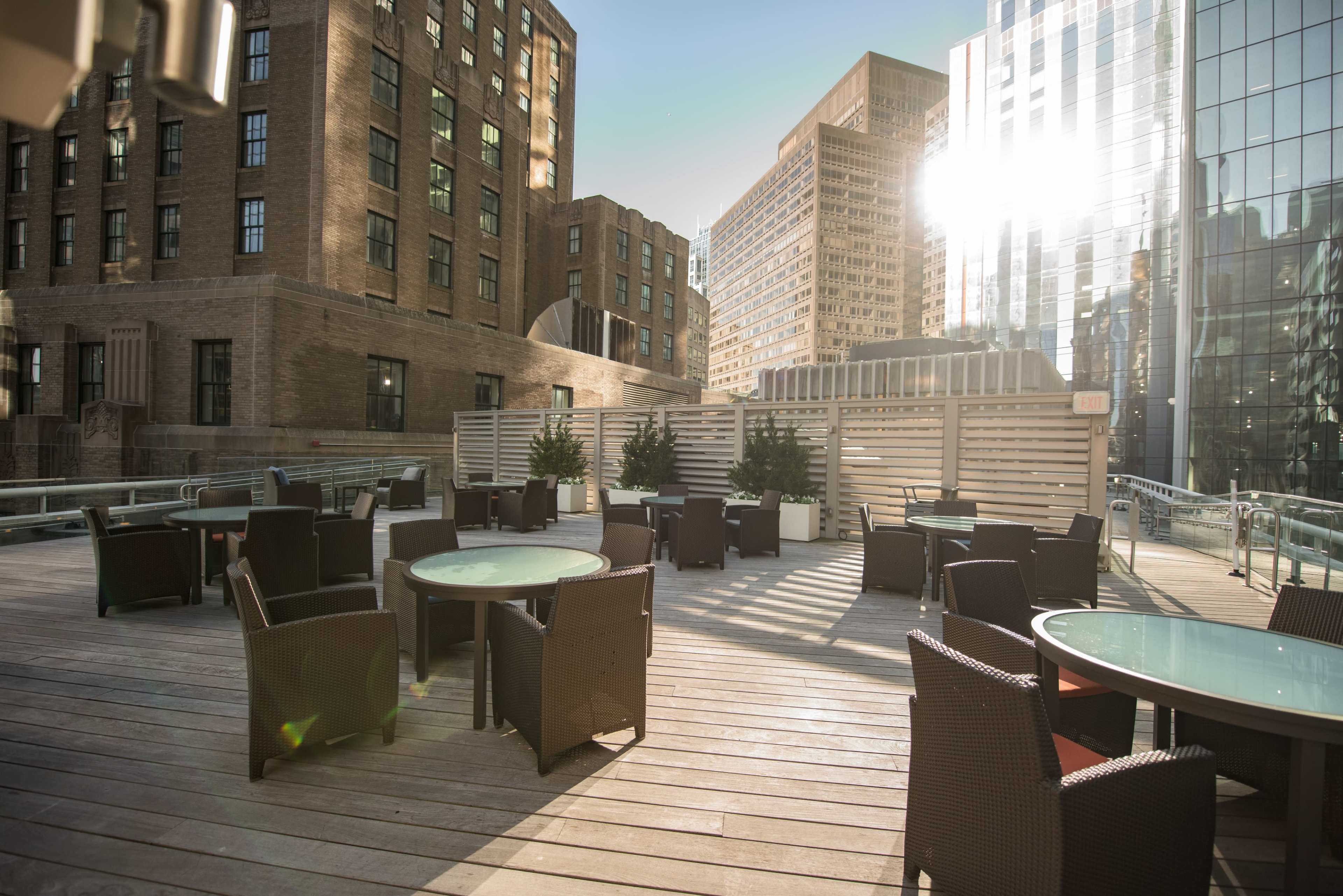 An outdoor patio area with wicker furniture and tables, surrounded by tall buildings reflecting sunlight.