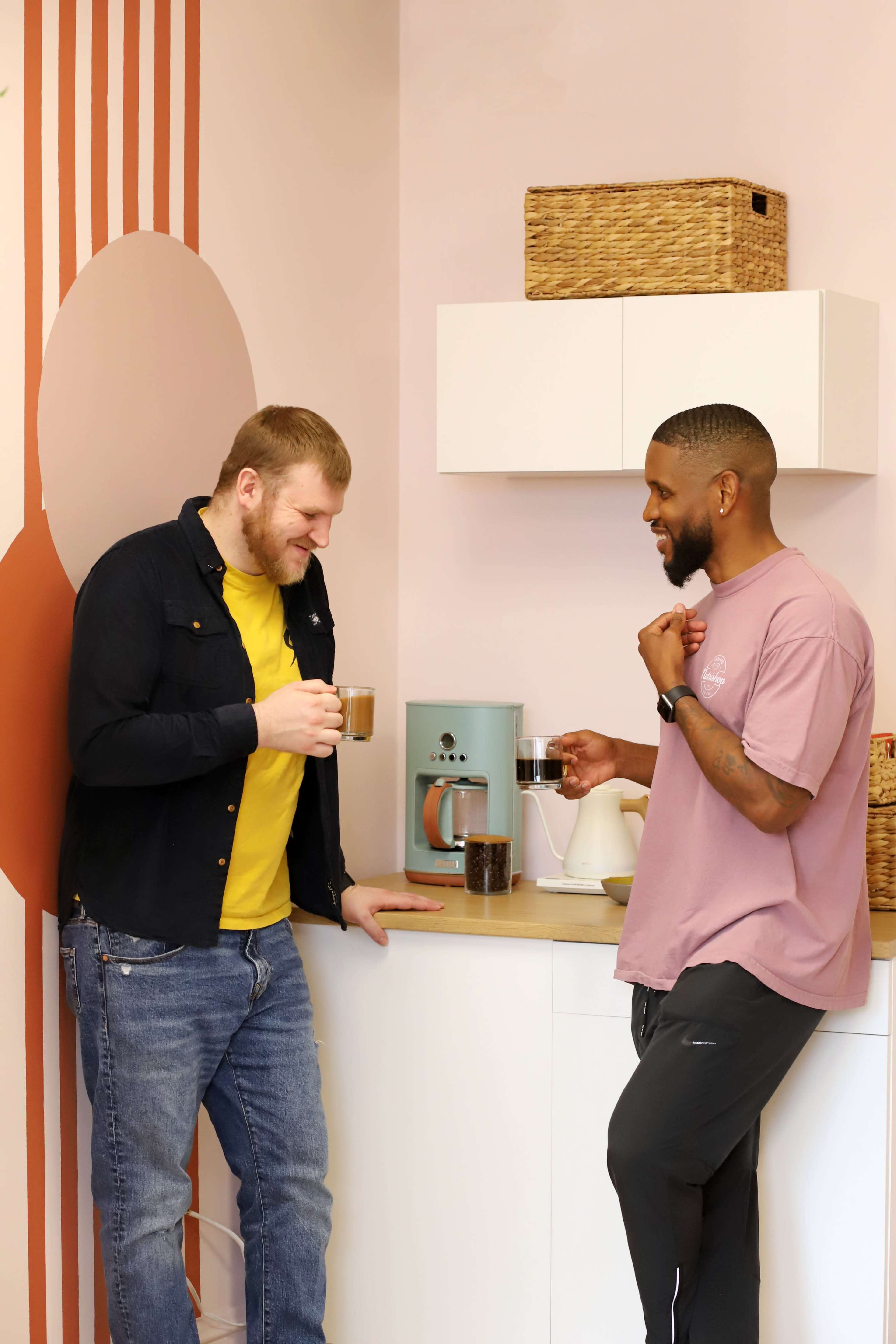 Two men stand in a kitchen, one holding a cup of coffee while the other holds a glass, facing each other near a coffee maker and containers.