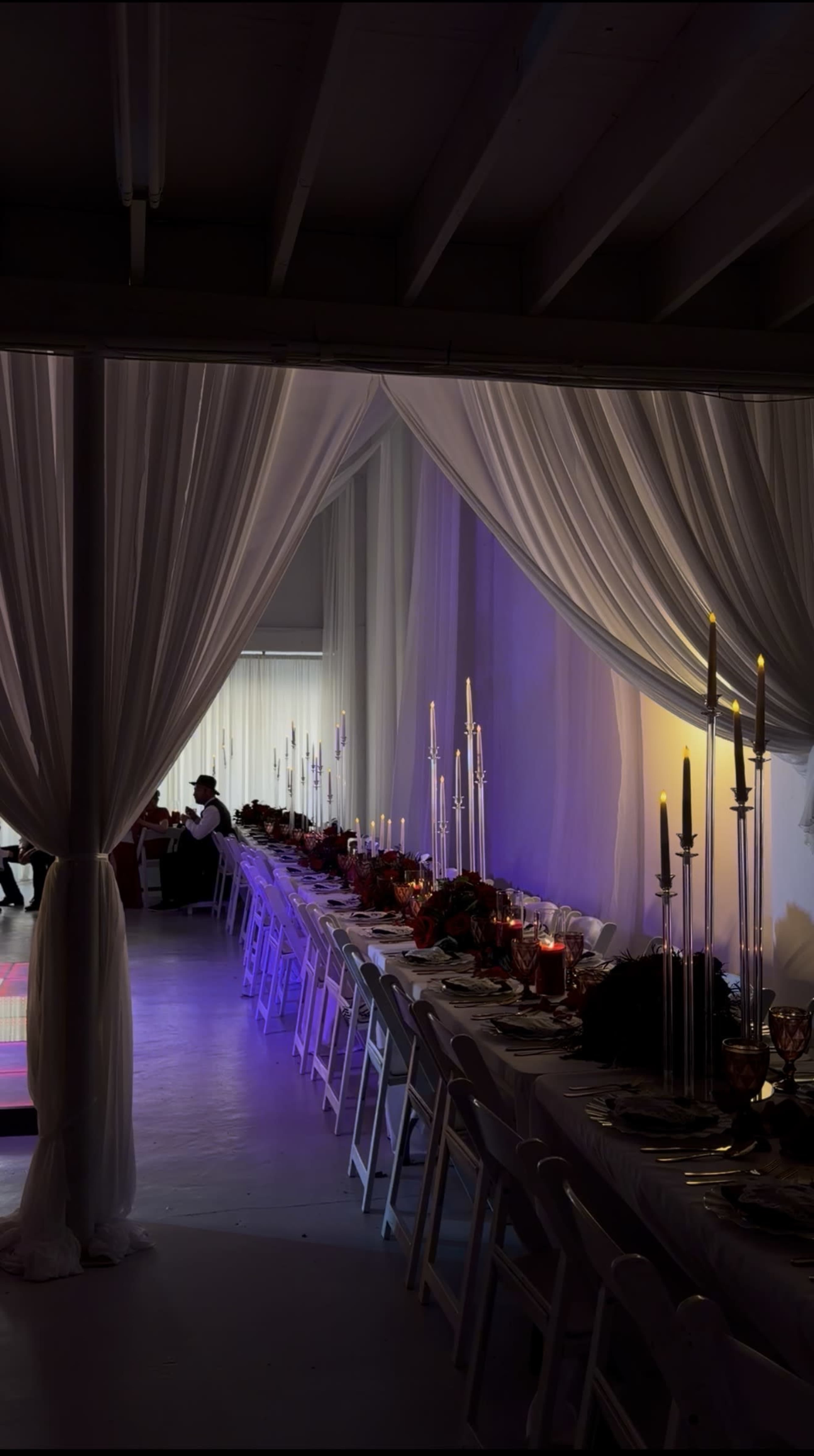 A long dining table is set with elegant tableware and candles, partially obscured by draped fabric in a softly lit venue.