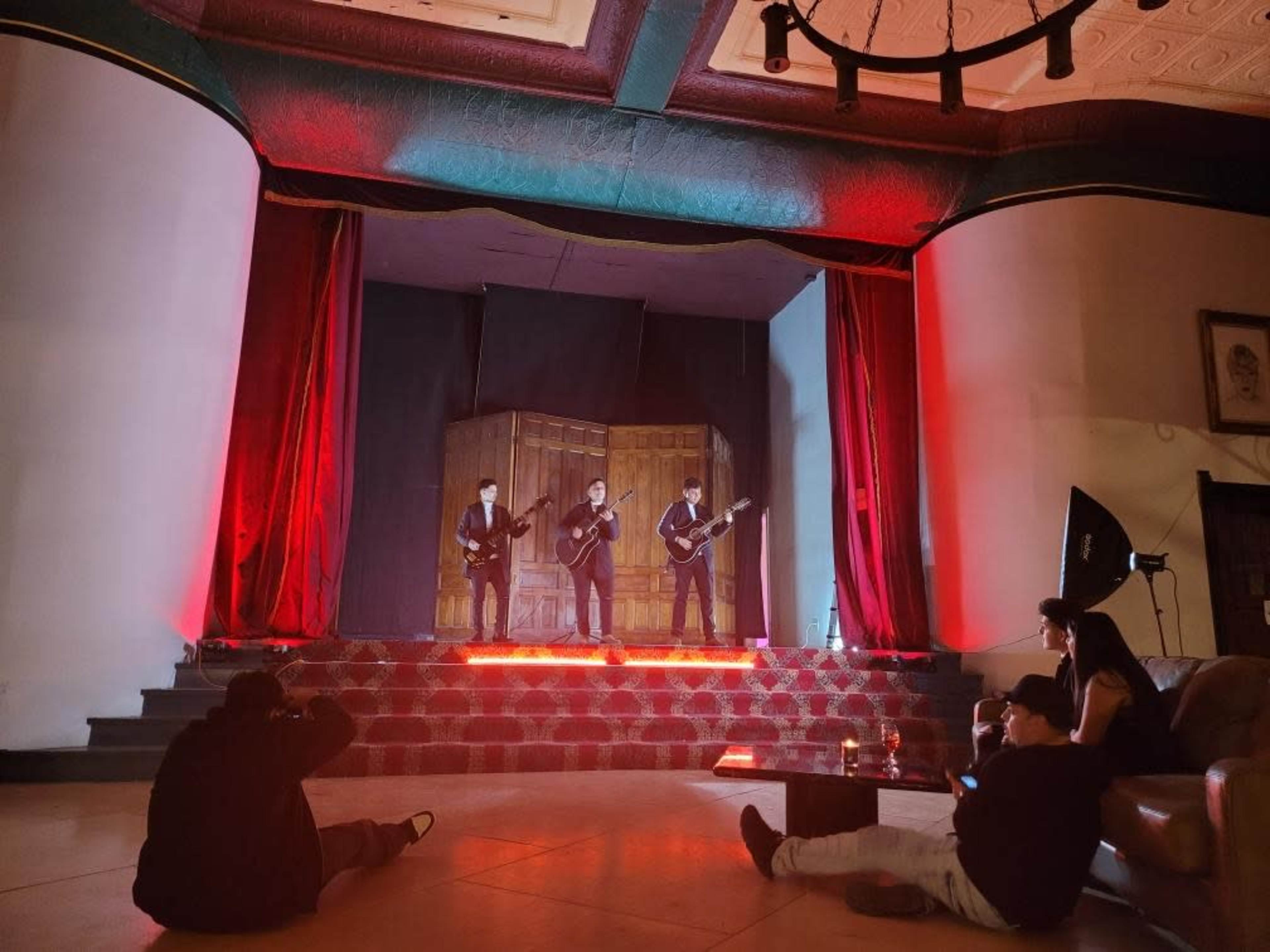 Three musicians perform on a stage with red lighting, while an audience sits on the floor and a couch in a well-lit venue.