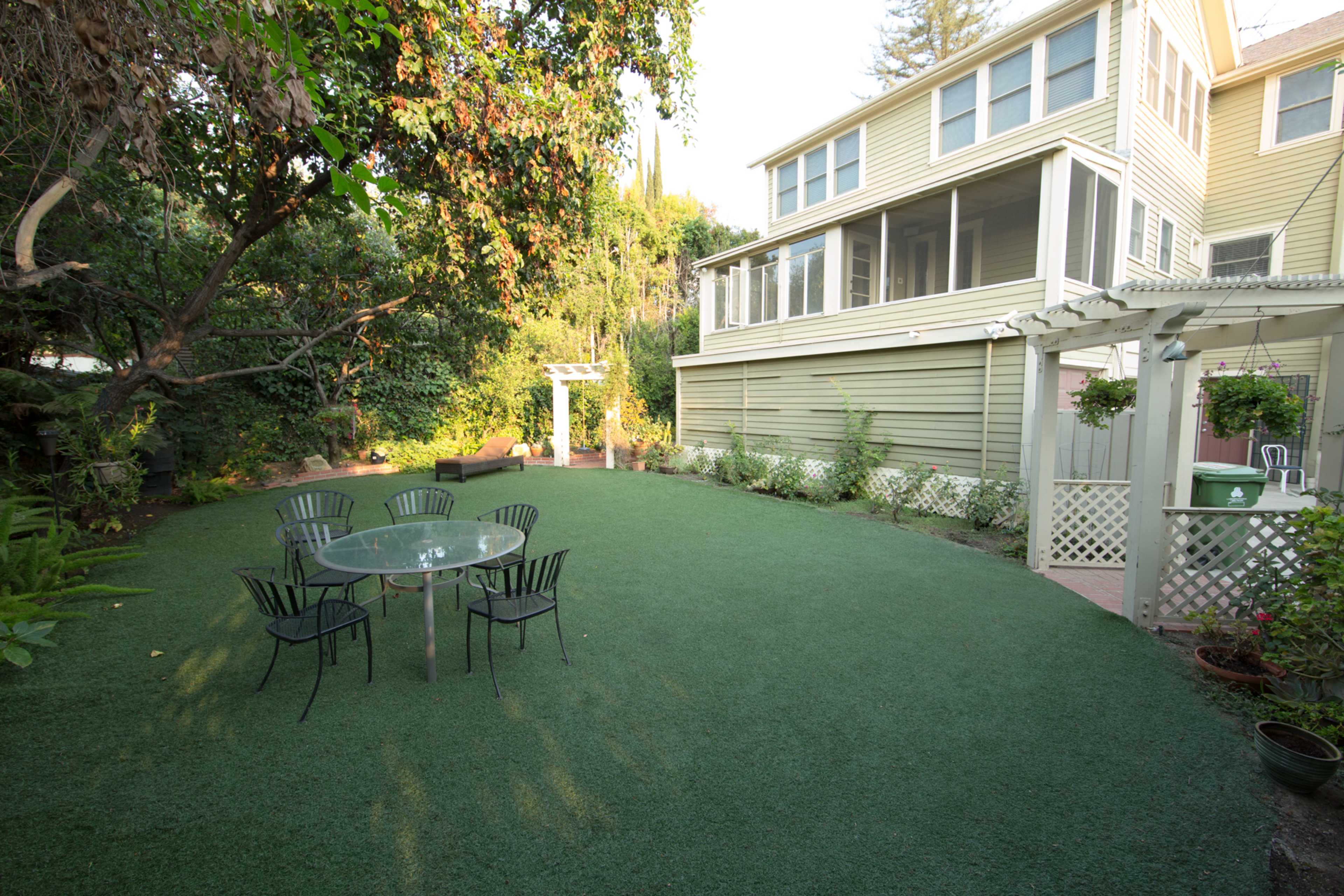 A backyard with artificial grass, featuring a glass table surrounded by black chairs and a multi-story house in the background.